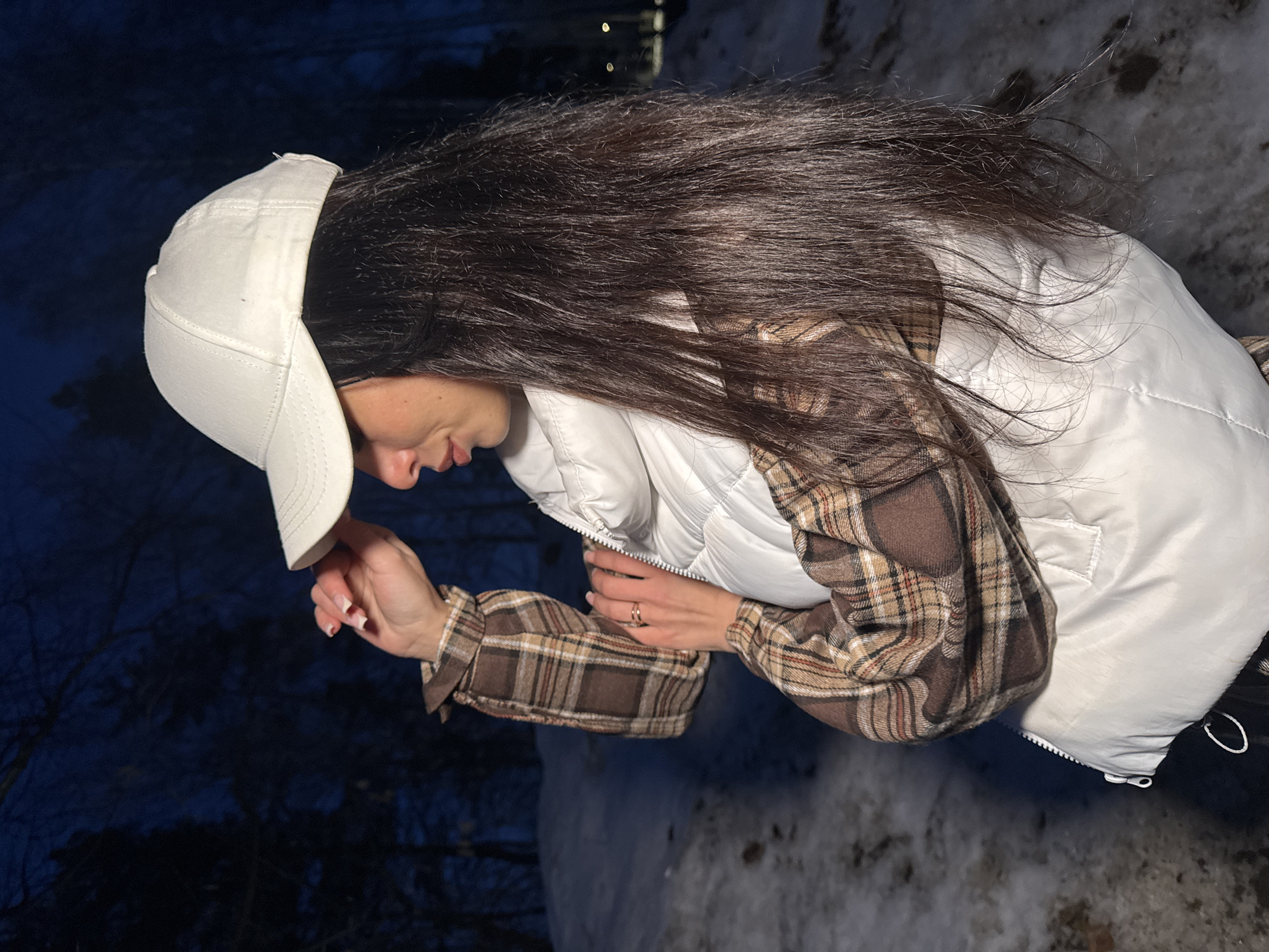 A woman wearing a white baseball cap, a white puffy vest, and a brown plaid shirt stands in an outdoor location. The background appears to be a snow-covered area, possibly at night or in low light. It may be in the city of Chaikovsky, Russia. The woman is looking down, possibly at something in her hand.