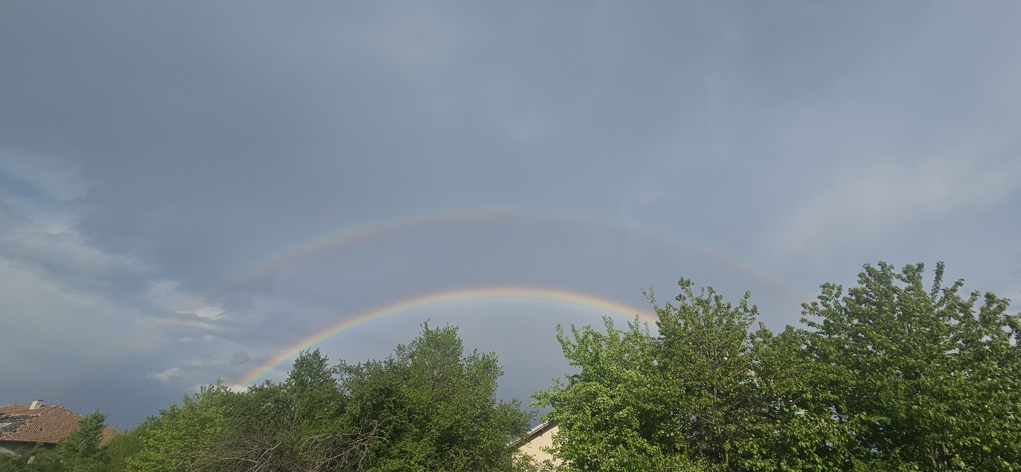 The image shows a double rainbow in the sky above green trees. Part of a building with a red tile roof is visible on the left. The sky is cloudy. The location is possibly near Gutsal, Bulgaria.