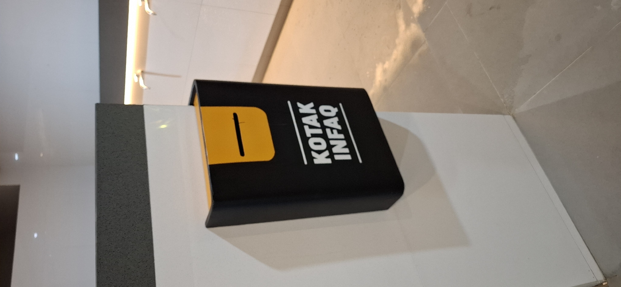 A black donation box with a yellow slot is mounted on a white wall. The text "KOTAK INFAQ" is printed in white on the box. The background shows a tiled floor and a white tiled wall with two faucets. The scene appears to be in a public restroom or a similar facility. The lighting is warm and comes from above. The overall environment is clean and modern. It is unclear if this image was taken in Ngemplak, Indonesia.