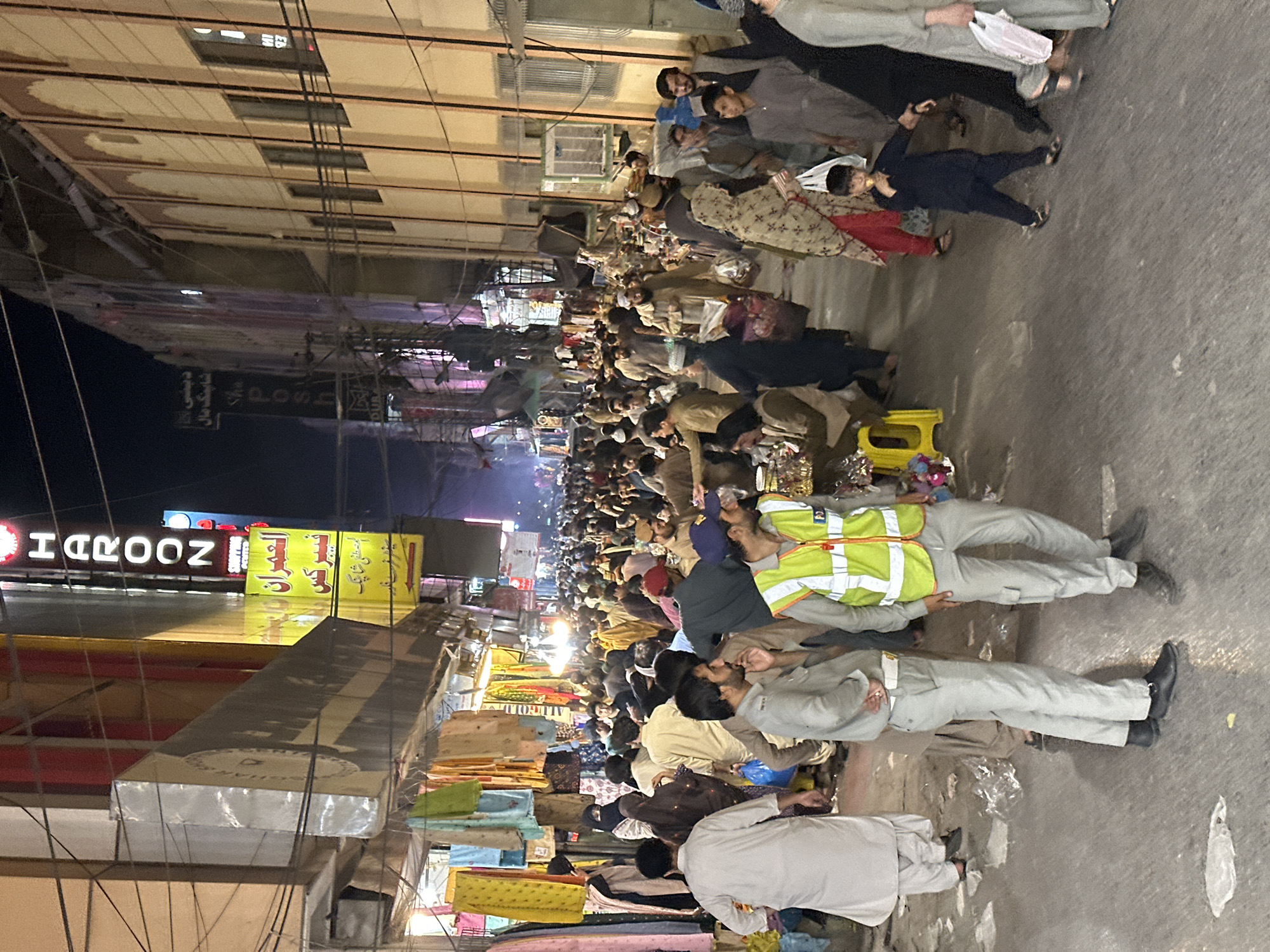 The image depicts a busy street scene in Quetta City Tehsil, Pakistan. A large crowd of people fills the street, with many seated or squatting on the ground. Some individuals are wearing traditional Pakistani clothing. Stalls and shops line the sides of the street, displaying merchandise such as clothing. There is at least one neon sign with the word "HAROON". The street is paved, and the lighting suggests it is nighttime. Some individuals are wearing vests, some of whom may be involved in security.