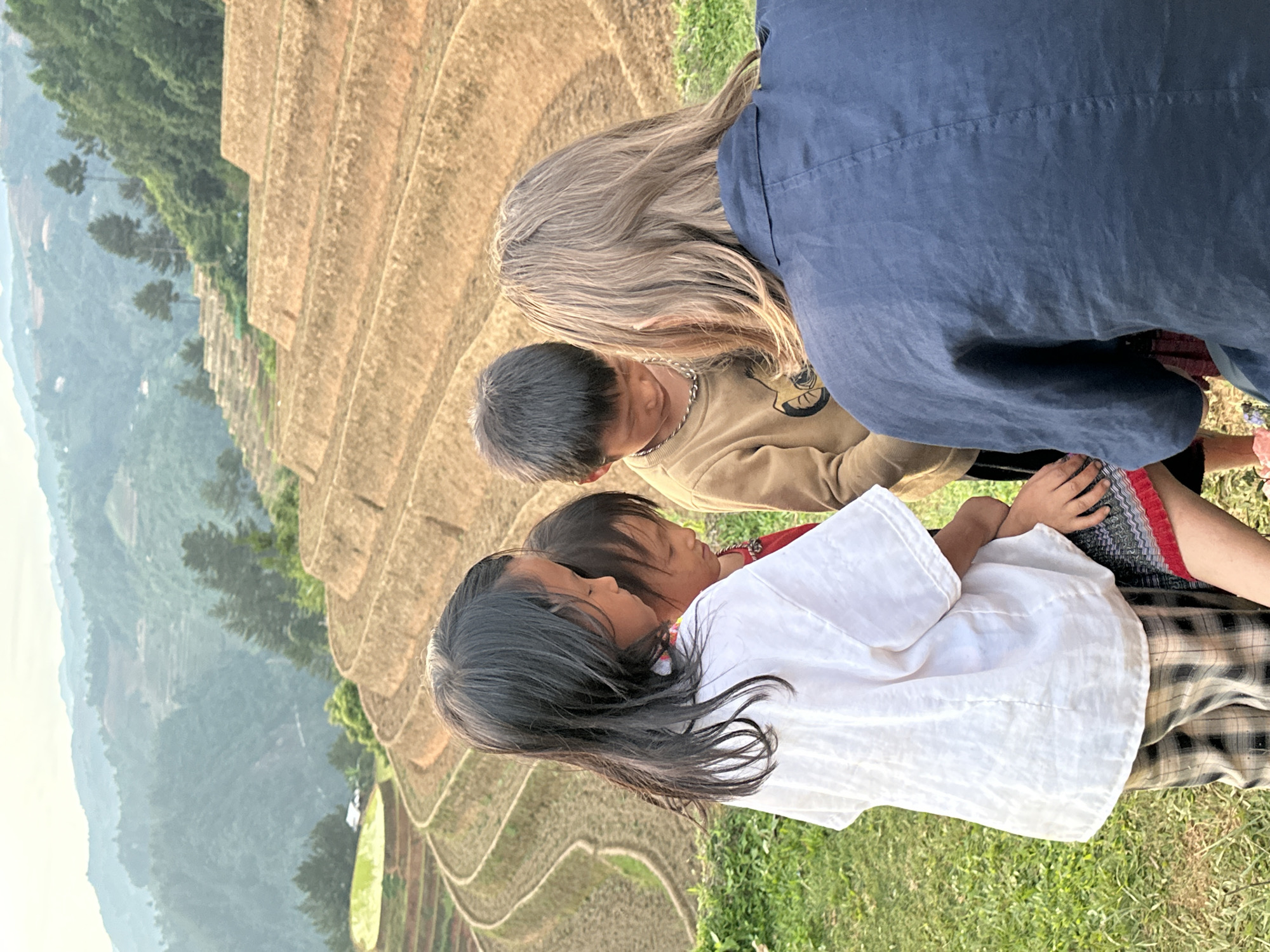This image captures an intimate moment amidst the breathtaking landscape of Mù Cang Chải, Vietnam. In the foreground, four individuals are closely gathered on a patch of green grass. An adult, viewed from behind, has long blonde or light brown hair and is wearing a dark blue top, leaning over the group. Below the adult, a child with short dark hair and a light brown long-sleeved shirt is visible. Prominently featured is another child with long, dark hair wearing a white shirt, their head gently resting against a third child, who is partially obscured but appears to be wearing red and a plaid garment. The close proximity and interaction among the children suggest a tender or observant moment.
The setting is dominated by the iconic terraced rice fields of Mù Cang Chải, which form a magnificent backdrop. These golden-brown terraces are intricately carved into the hillsides, stretching across the mid-ground and receding into the distance. Beyond the terraces, lush green mountains fade into a soft, hazy horizon under a bright sky. The warm, diffused lighting suggests either early morning or late afternoon. The scene conveys a sense of peacefulness and connection between the people and the natural environment. There is no visible text in the image.