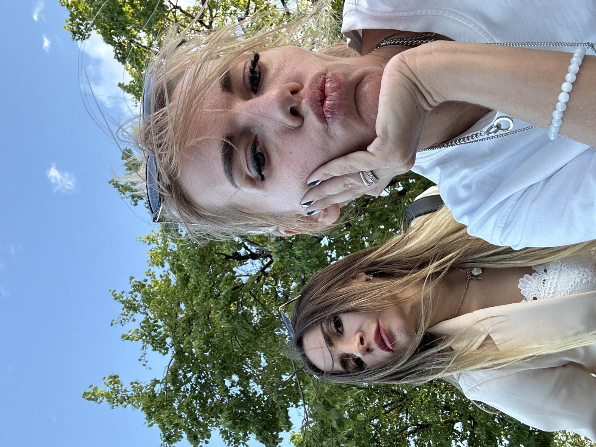 Two women are captured in a selfie outdoors. The woman in the foreground, with blonde hair styled in a messy bun and sunglasses perched on her head, is puckering her lips and resting her chin on her hand. She wears a white t-shirt adorned with a silver chain necklace and a bracelet of white beads. The woman behind her, with long brown hair, gazes directly at the camera with a serious expression. She is wearing a cream-colored blazer over a white lace top and a delicate necklace. Lush green trees and a clear blue sky form the backdrop, suggesting an urban park setting. The lighting indicates it is daytime. While the image does not provide explicit markers for the location, the presence of mature trees and the overall ambiance are consistent with many green spaces found within cities like Saint Petersburg, Russia, such as Yelagin Island or the Summer Garden.