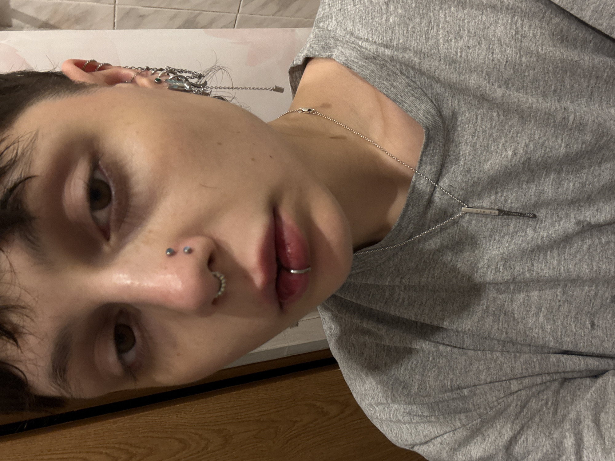 A person with multiple piercings, including in the nose, lip, and ear, is wearing a gray t-shirt and a silver necklace. The background appears to be a bathroom with tiled walls. The location is unknown.