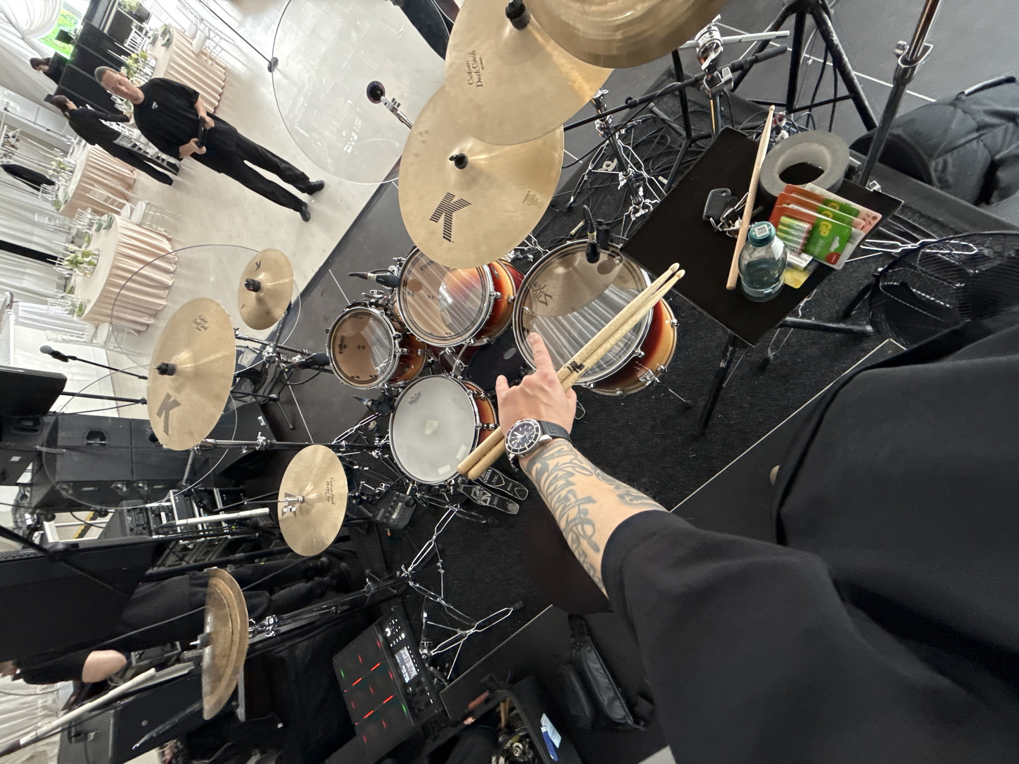 The image shows a drum set from a high-angle perspective. The drums are positioned on a black rug or platform. A person wearing a black shirt and a wristwatch is holding drumsticks over the drum set. Their arm is tattooed. There's a black table nearby with a water bottle, tape, and other items. Other people are visible in the background, standing near tables in what appears to be a venue or event space. The location is possibly Shchekutino, Russia, though this cannot be confirmed solely from the image content.