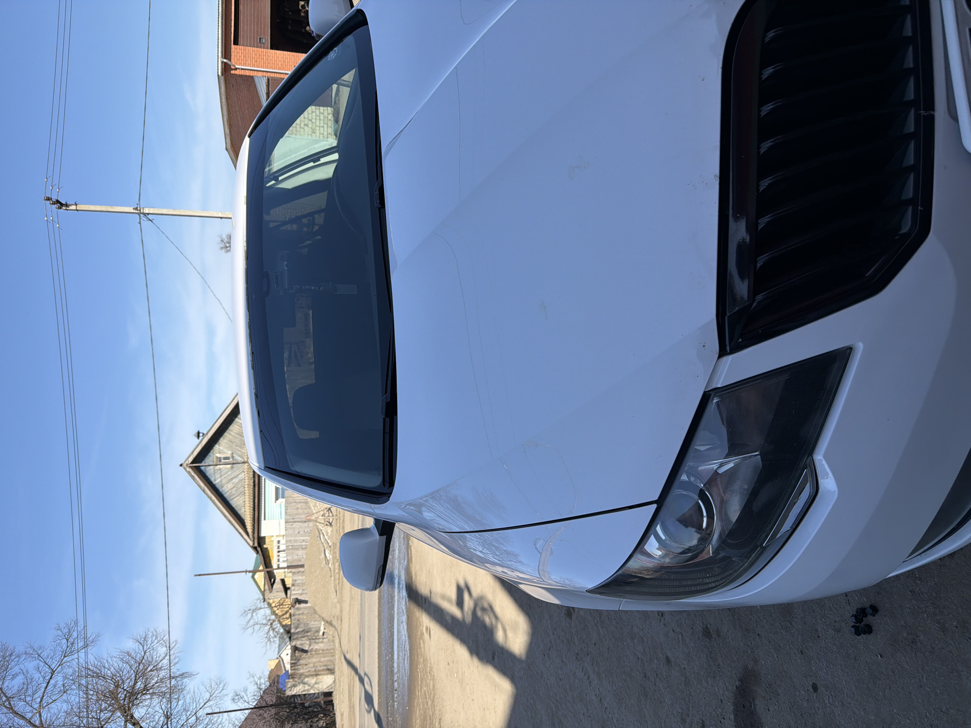 A close-up, slightly distorted photograph features the front of a white car parked on a paved surface, likely a street or parking area. To the left of the car, the reflection in the windshield shows a view of houses, telephone poles, and trees under a clear blue sky. The vehicle is positioned close to the right edge of the image, with its black grill prominently displayed. On the ground near the front of the car are several small, dark objects, possibly nuts or bolts. The location is believed to be in Chaikovsky, Russia, based on the buildings and environment visible in the reflection.