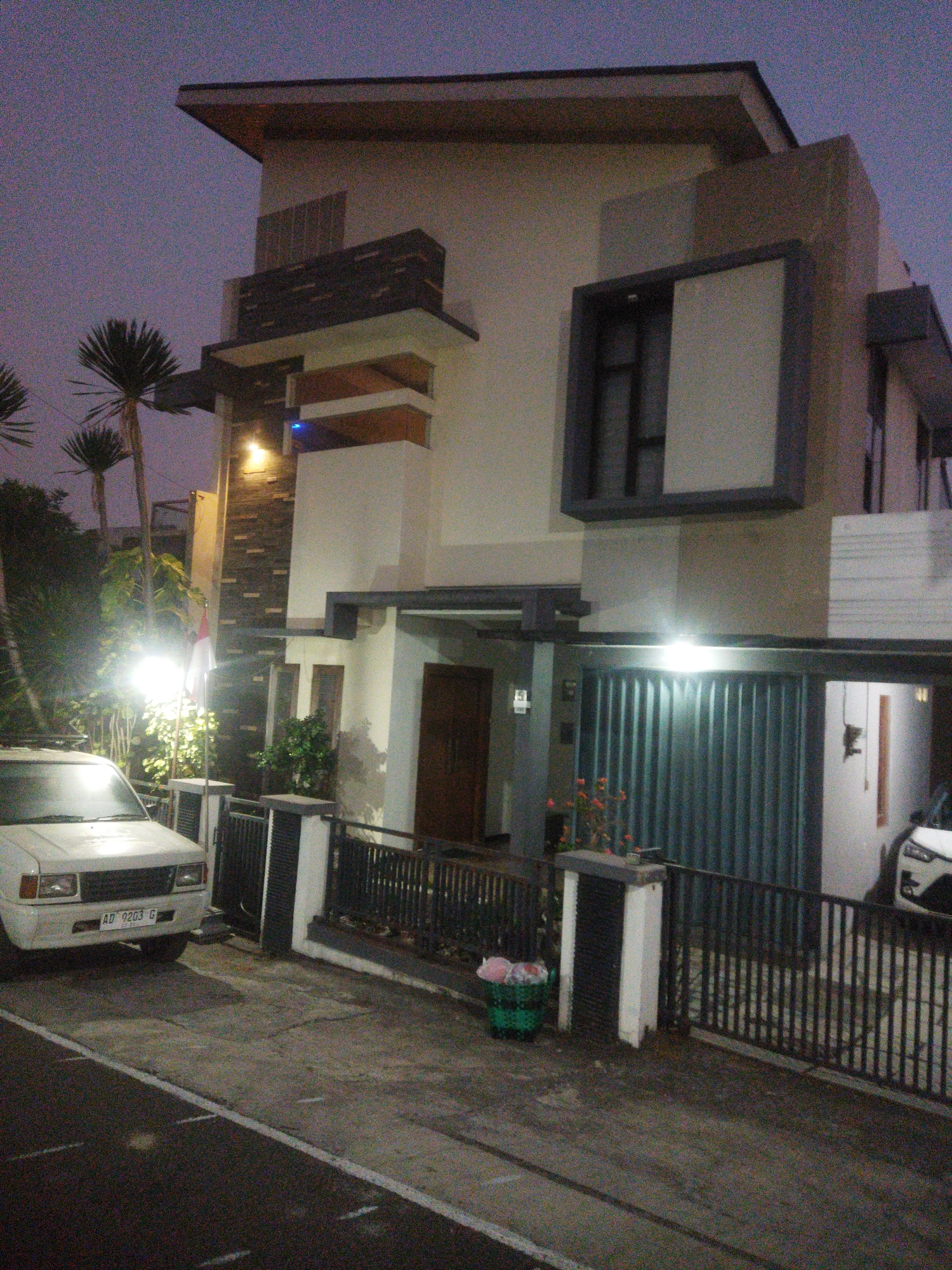 The image depicts a modern two-story house in the city of Wonogiri, Indonesia. The house features a combination of white and beige walls with dark accents around the windows and roofline. A dark metal gate guards the entrance to the property, which includes a covered carport. A white vehicle is parked in front of the house. The scene is illuminated by artificial lights, suggesting it is dusk or nighttime. The environment appears to be a residential street with a paved road in the foreground. Palm trees and other vegetation are visible on the property. There are no people visible in the image.