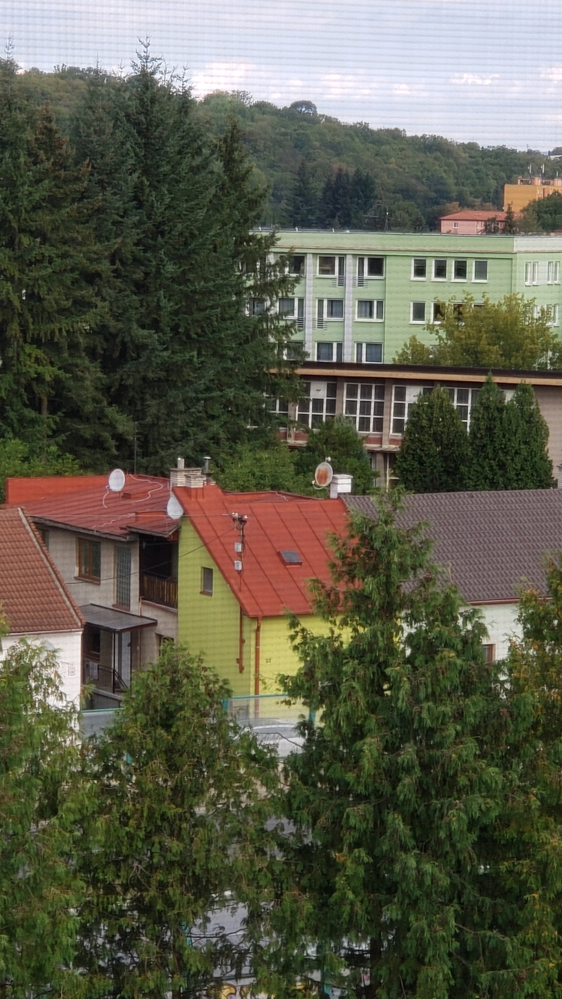 The image depicts a residential area with a cluster of houses and apartment buildings surrounded by lush greenery and trees. The foreground is dominated by the roofs of several houses, characterized by their red tiles and varying architectural styles. One house in particular stands out with its lime green exterior, featuring a prominent red roof and a chimney. To the right, another house has a brown, textured roof.
In the mid-ground, several large evergreen trees with dense foliage are visible, partially obscuring some of the buildings. Behind these, a light green, multi-story apartment building with numerous windows is prominent. Further in the background, a gentle, tree-covered hillside rises, with a glimpse of a yellow building and a smaller pink building on its slope. The sky above is overcast, with soft clouds.
The scene appears to be a tranquil, suburban setting. There are no people visible in the image. The presence of satellite dishes on some of the house roofs suggests modern amenities within these residential structures. The overall impression is one of a peaceful, established neighborhood.
The image captures a view of buildings and trees that are consistent with the urban landscape of Prešov, Slovakia, a city known for its blend of residential areas, green spaces, and rolling hills. The architectural styles of the houses and the presence of apartment blocks are typical of many European cities. The dense greenery and the hillside in the background are also characteristic features of the Prešov region.
