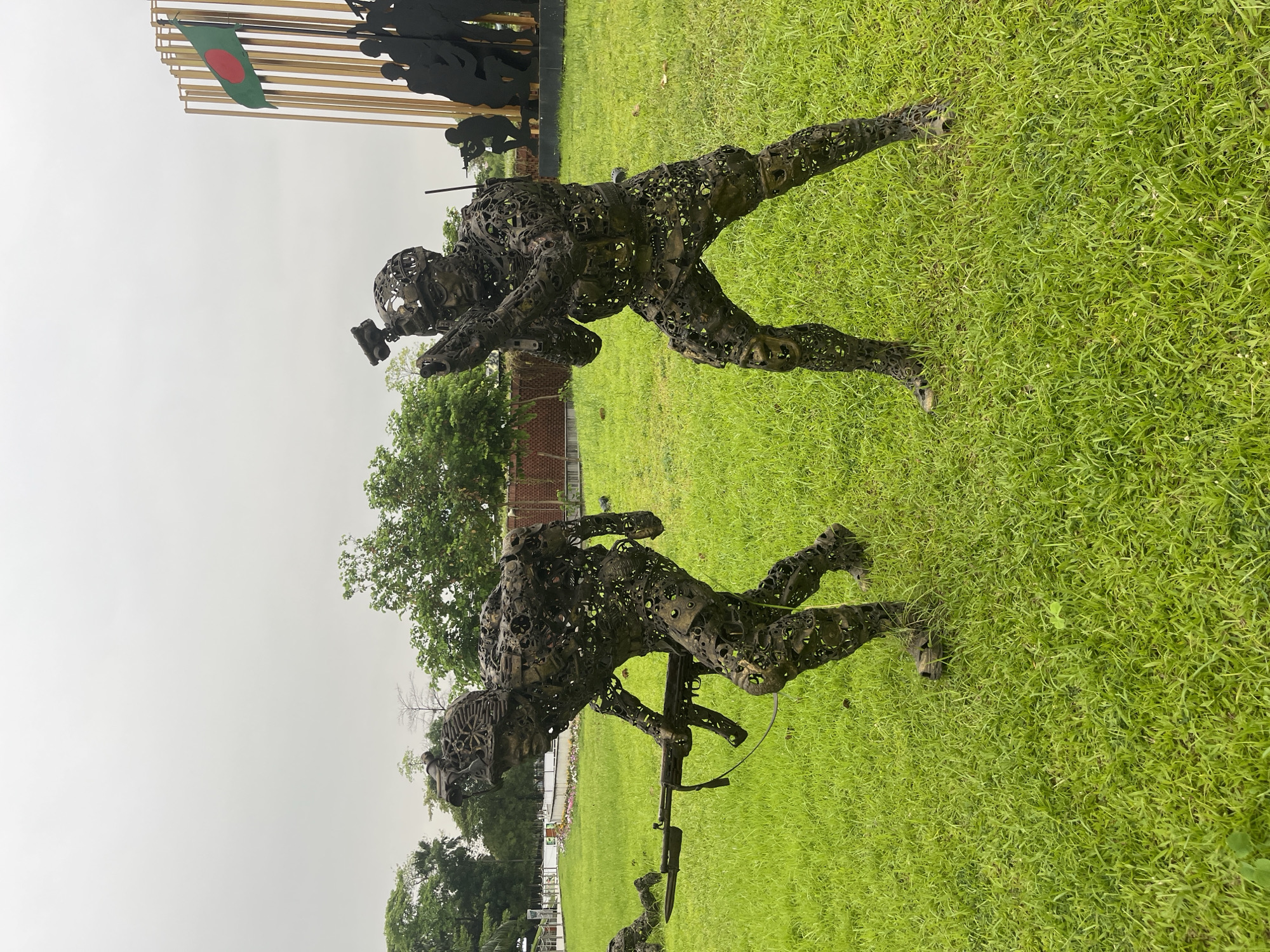 The image shows a memorial in Dhaka, Bangladesh featuring two metal soldier sculptures positioned on a lawn. The sculptures are depicted in combat poses; one holding a firearm and the other with an object raised above. In the background, the national flag of Bangladesh is visible along with silhouettes of additional figures. The environment is a grassy area, seemingly a park. The sky is overcast.
