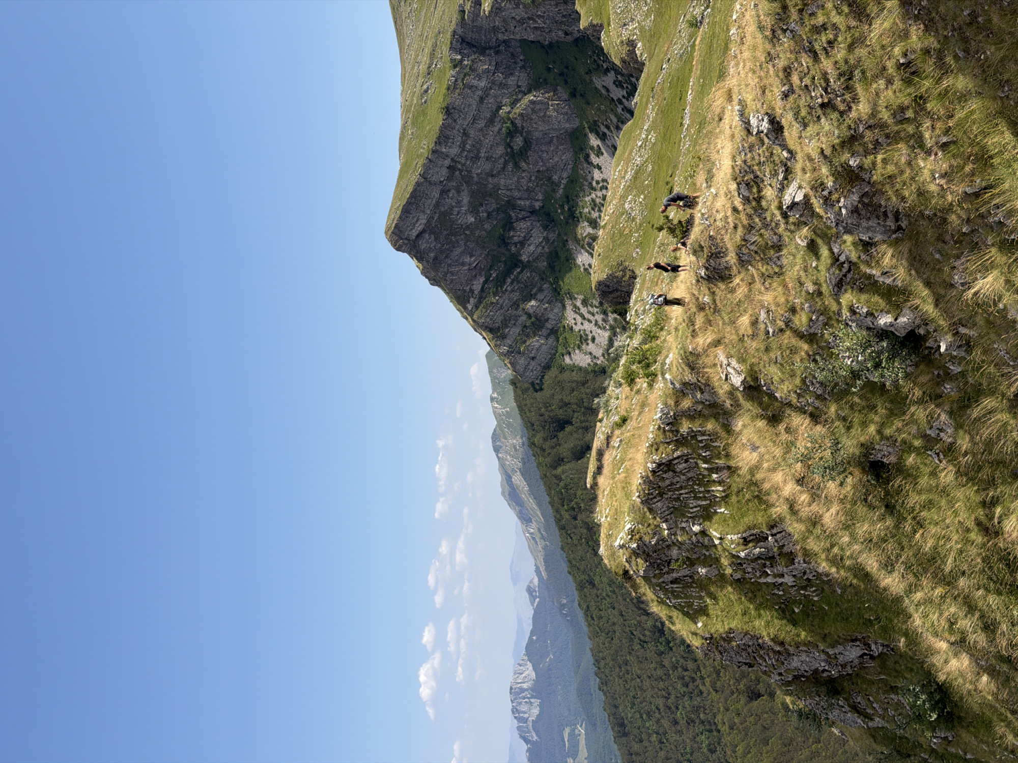 Four individuals are observed standing on a grassy, rocky mountain ridge. The person furthest left is wearing a backpack and dark clothing. Next to them, another person in dark clothing stands. To their right, a third individual in dark clothing is seated, and a fourth person in dark clothing is bending over, possibly interacting with the seated individual or an object on the ground. The terrain is characterized by rugged, rocky outcrops and slopes covered in dry, yellowed grass. In the background, a dense forest of green trees covers a mountainside, receding into the distance where layered mountain ranges are visible under a clear, light blue sky with scattered white clouds. The scene depicts an outdoor activity, likely hiking or exploring, in a mountainous environment.