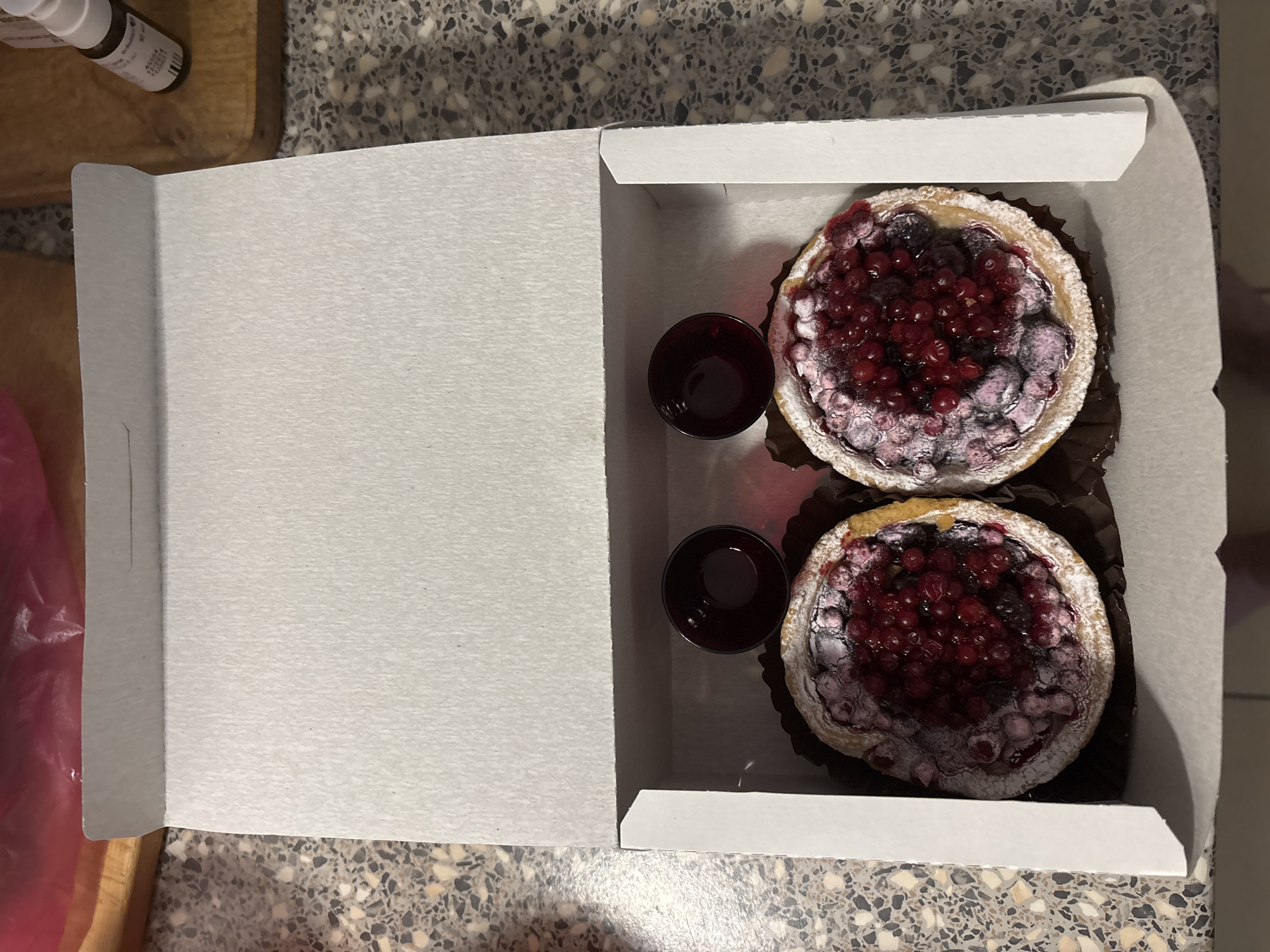 A top-down shot captures an open white cardboard box on a speckled, grey countertop, with a portion of the box open and filled with food. Two small, round tarts filled with red berries, possibly red currants or cranberries, sit within the box, appearing to be dusted with powdered sugar. Two small cups filled with dark liquid are positioned next to the tarts. The image does not provide explicit details suggesting the location is Saint Petersburg, Russia.