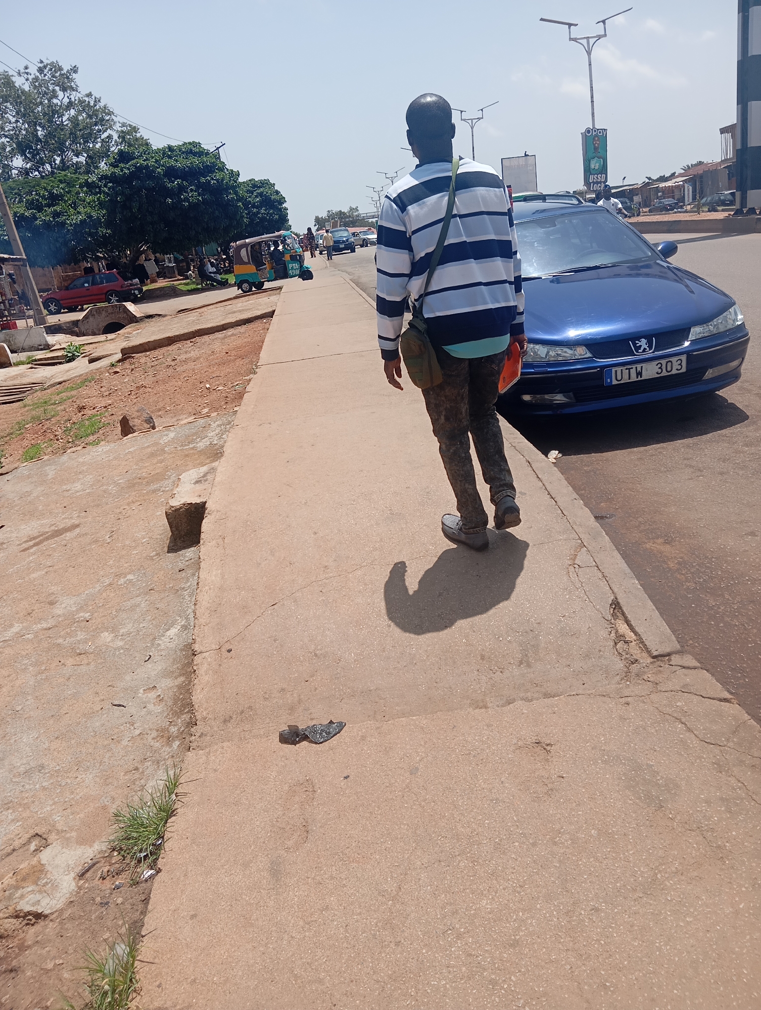 A man walks along a concrete sidewalk next to a blue Peugeot sedan on a sunny day in Jos, Nigeria. The man is wearing a striped long-sleeved shirt and camouflage pants, with a bag slung over his shoulder. The car has a license plate that reads "UTW 303". In the background, a three-wheeled taxi is visible, along with trees, other vehicles, and buildings. The sidewalk is cracked and worn, and the surrounding area appears to be a commercial or roadside setting.