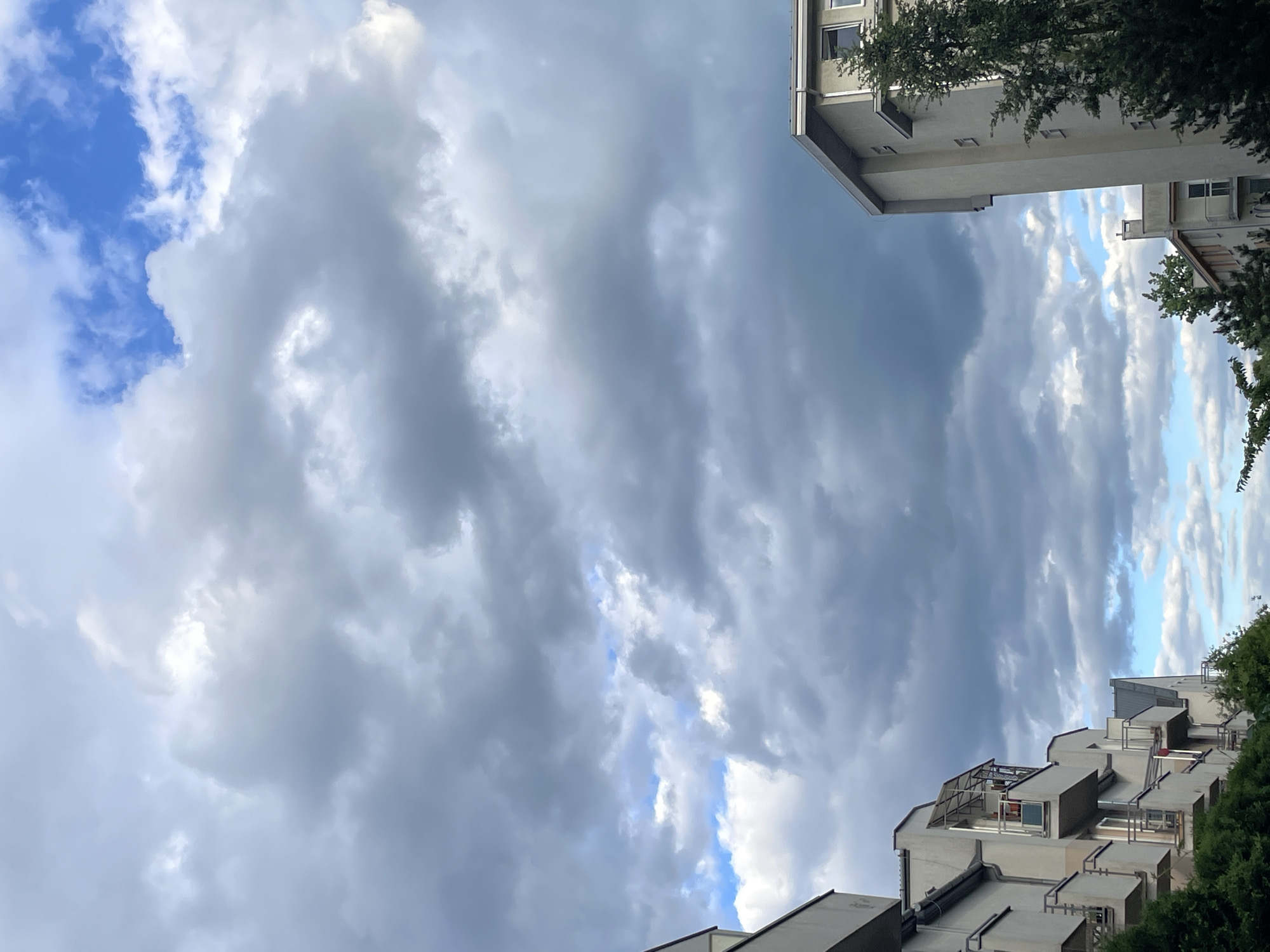 The image shows a view of the sky with heavy cloud cover, partially obscuring areas of blue sky. The perspective is from below, looking upwards. In the corners of the image, portions of buildings are visible, suggesting an urban setting. There is some greenery, possibly trees, also visible along the edges of the buildings. The location is Unknown, Unknown.