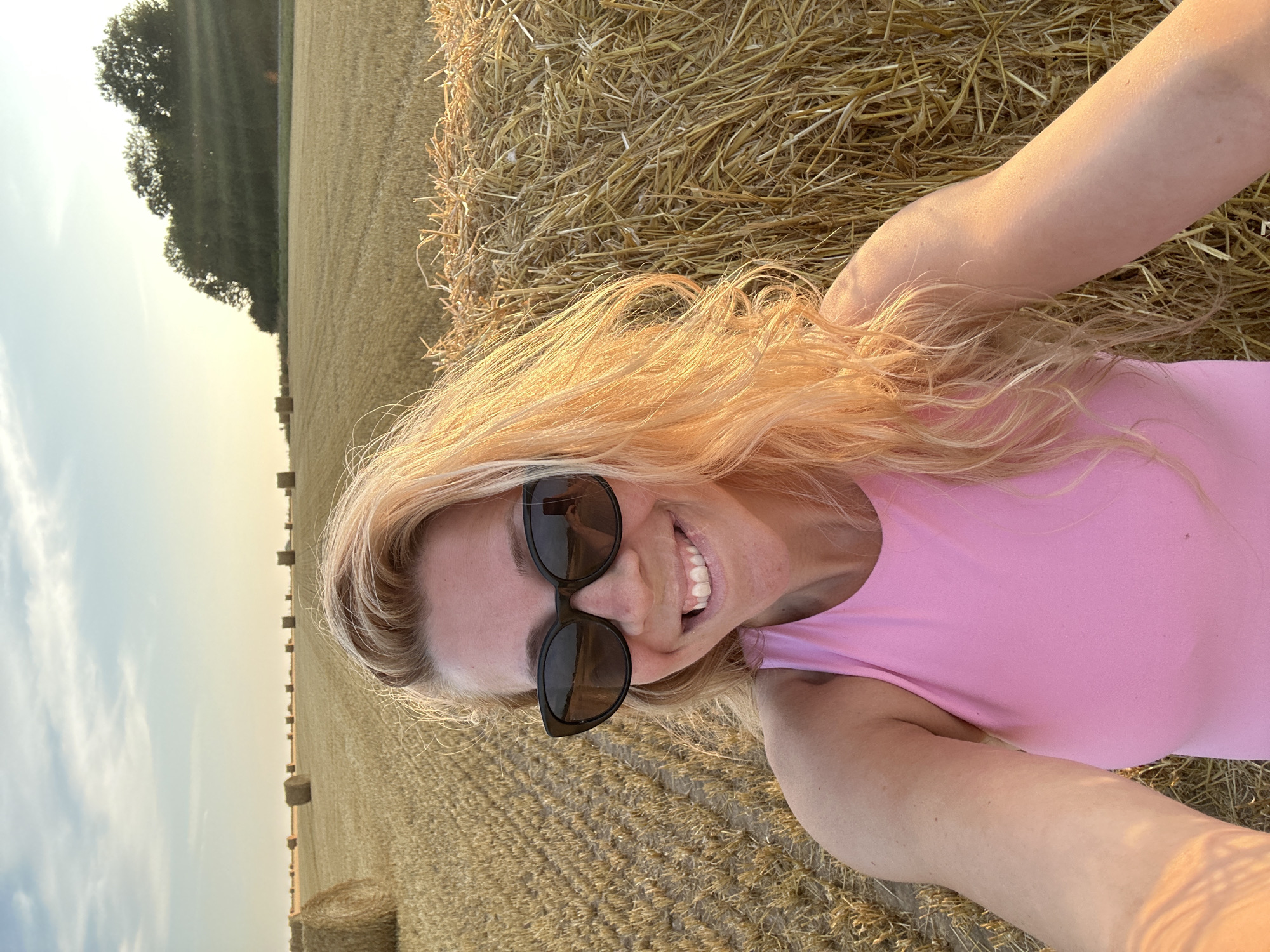 The image is a selfie taken outdoors in a field of hay bales, likely in Trouville-Alliquerville, France. A woman with blonde hair and sunglasses is smiling at the camera, wearing a pink top. She is positioned in front of a large hay bale. The background shows a field with more hay bales stretching into the distance under a hazy sky with the sun setting.
