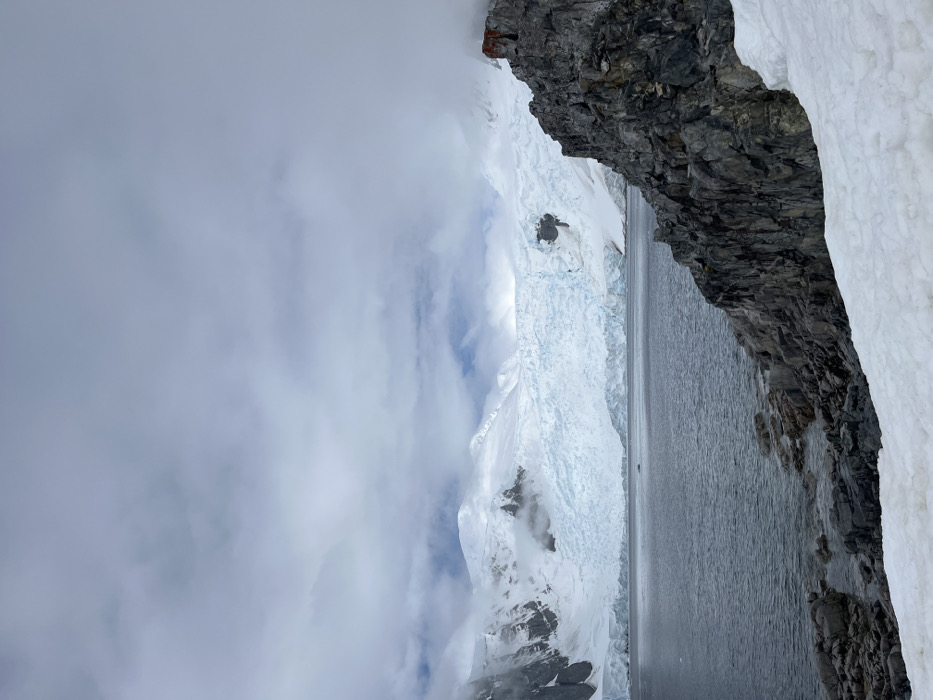 A vertical shot showcases a stark landscape. A jagged, dark rock formation with snow accents on the right frames a gray body of water. A vast glacier ascends on the opposite side, partially obscured by clouds. Snow-capped mountains rise beneath the glacier, disappearing into the overcast sky.