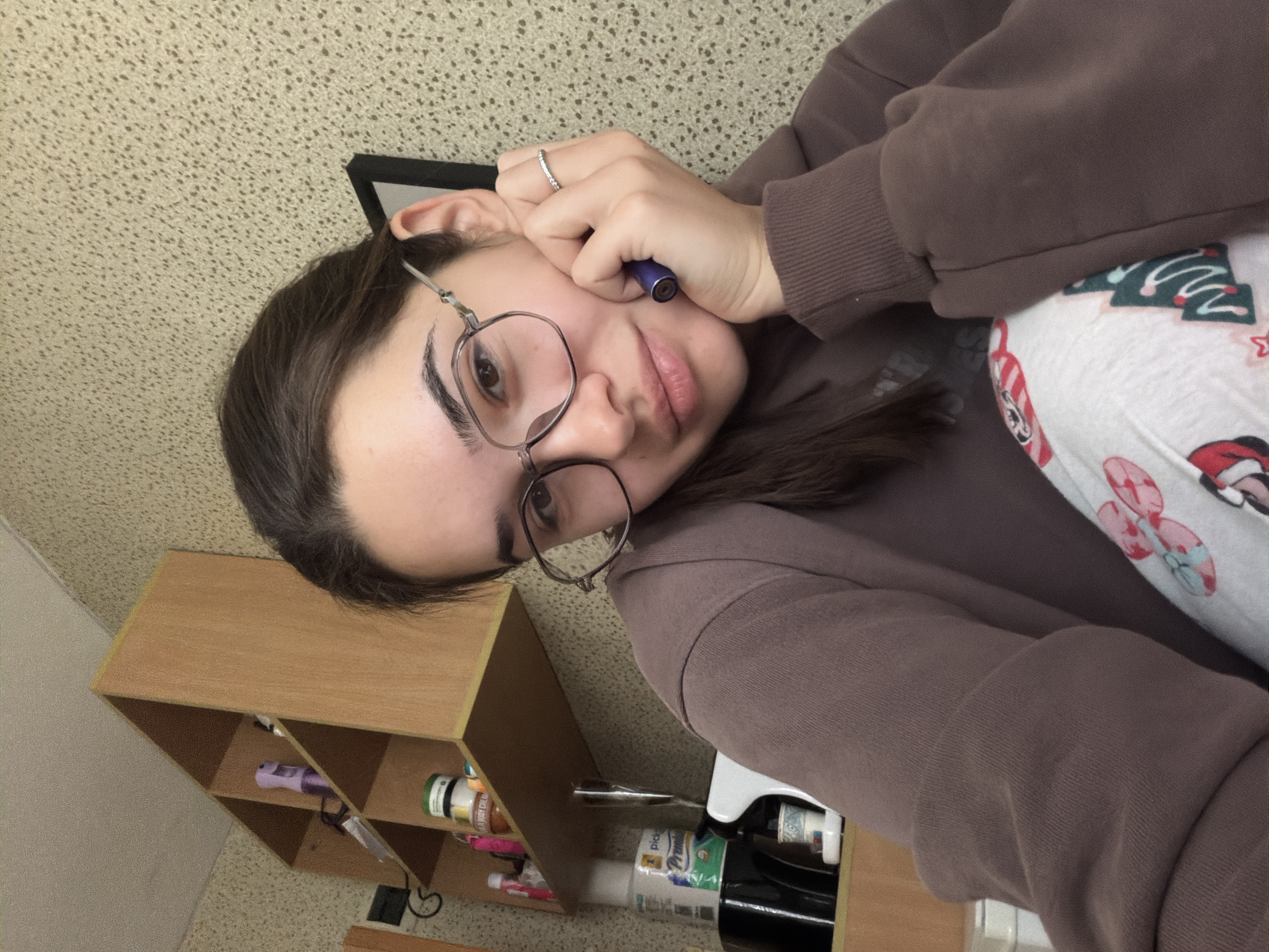 A casual self-portrait depicts a young woman indoors. She has dark hair, dark eyebrows, and is wearing round, metal-rimmed glasses. Her lips are slightly pursed, conveying a neutral or contemplative expression. She wears a dark brown, oversized hoodie with ribbed cuffs and has a silver-colored ring on the ring finger of her left hand. She is holding a small, dark blue cylindrical object, possibly a vape pen, near her cheek.
The background shows a textured, light-colored wall or ceiling in the upper portion of the image. To her right, part of a white garment with a festive pattern, including stylized green Christmas trees, red candy canes, and possibly Santa Claus heads, is visible, suggesting she might be wearing pajamas or covered by a holiday-themed blanket.
In the lower left, a light wooden shelving unit with multiple compartments is visible. Various items are stored on these shelves. One shelf contains a purple cylindrical object, possibly a hair styling tool. Another shelf holds several bottles and containers, including a white bottle with a green label that reads "body cream" and a partially visible brand name "Pick 'n Pay Premium." A dark glass bottle, possibly of wine or spirits, is also present on a lower shelf.
The image is captured from an overhead perspective and rotated approximately 90 degrees clockwise, making the subject appear on her side. There are no specific visual cues within the image to indicate the location as Sofia, Bulgaria. The setting appears to be a casual indoor space, possibly a bedroom or living area.