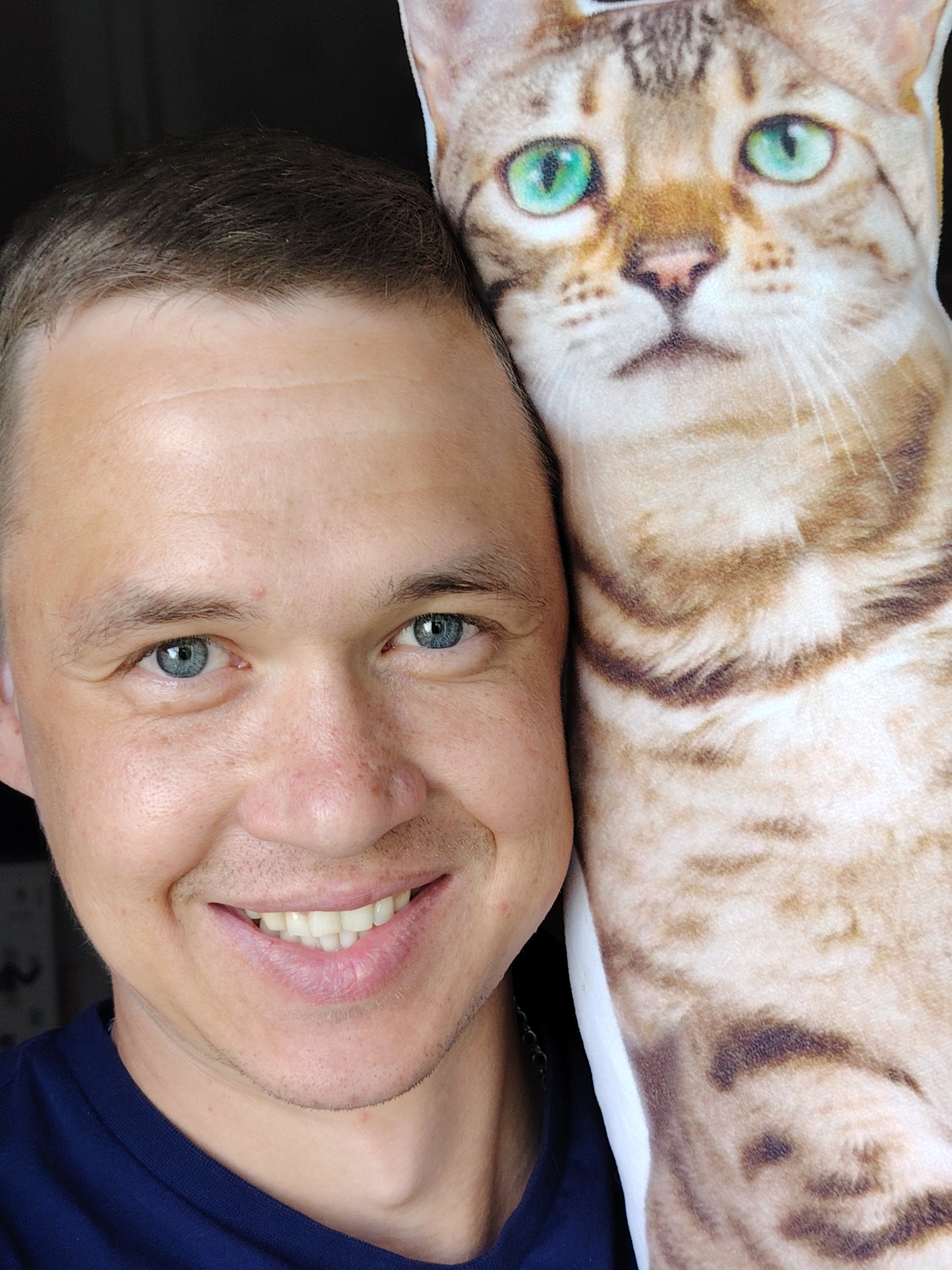 A close-up captures a smiling man with short, light-brown hair and blue eyes, positioned beside a large cat-shaped pillow. The pillow is printed with a realistic image of a light-brown tabby cat with bright green eyes. The man wears a dark-blue shirt. The location is not discernible from the provided image to determine if it is in Chaikovsky, Russia. No specific actions or interactions are observable beyond the man's smile and proximity to the pillow. The environment is generally neutral and dark.