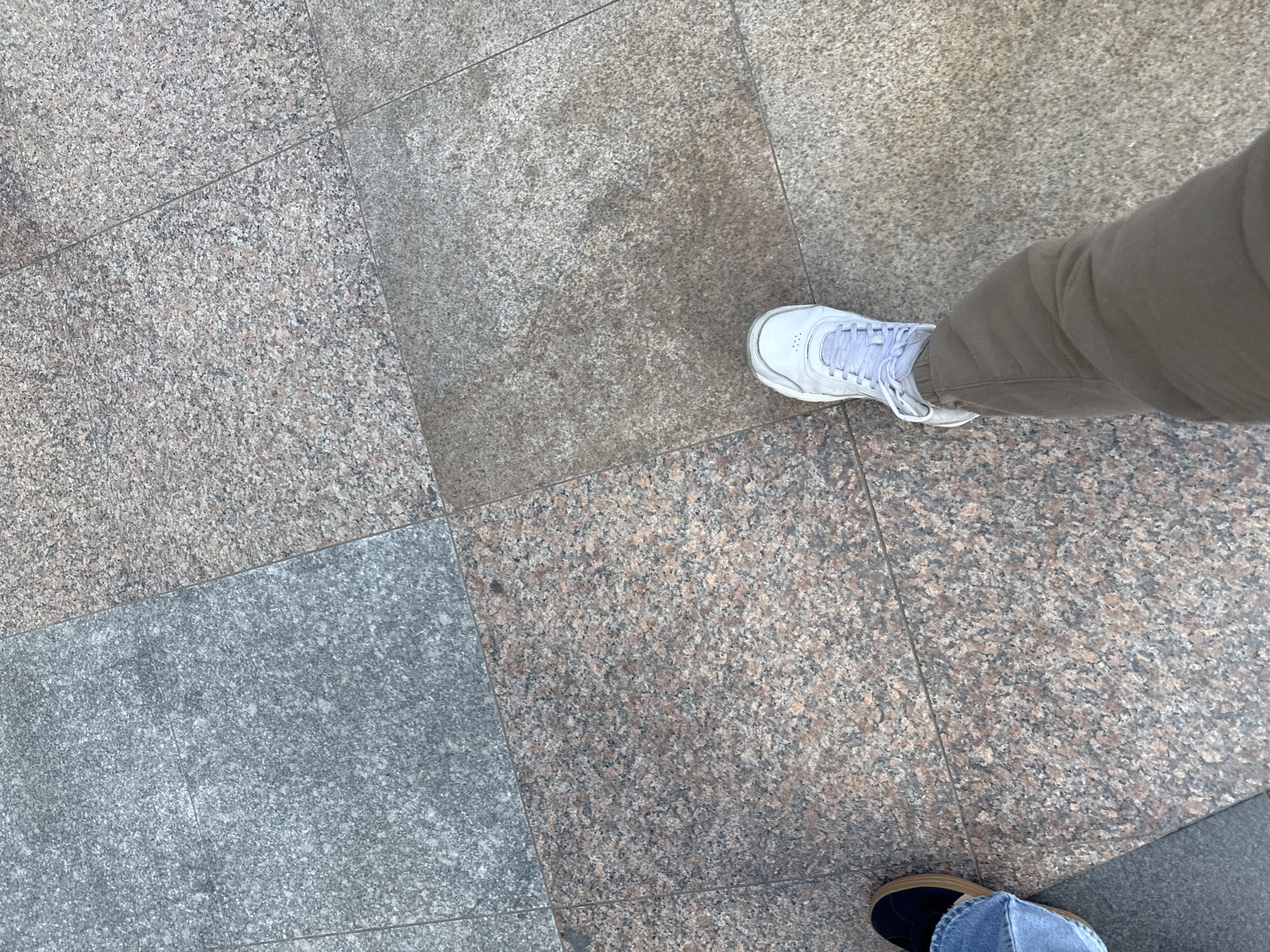 The image shows a ground-level view of tiled paving in Sofia, Bulgaria. The tiles are of a mottled granite-like material in varying shades of gray and reddish-brown. Two feet are visible. The right foot is wearing a white sneaker and a brown/khaki-colored trouser leg is visible. The left foot is wearing a blue denim pant leg and a dark-colored shoe. There are no visible interactions or actions. The environment consists solely of the tiled pavement.