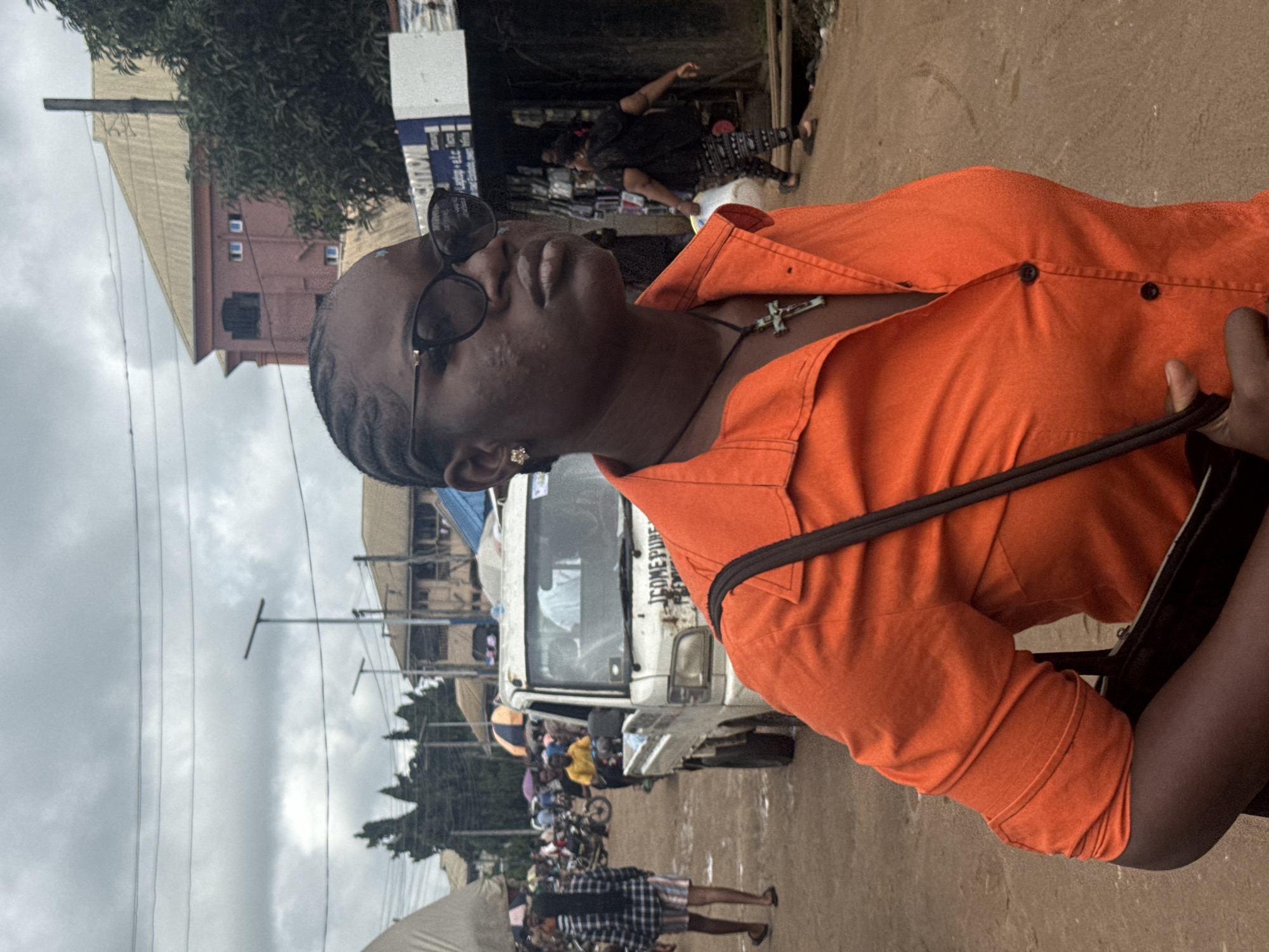 The image captures a medium shot of a young Black woman from the chest up, facing right, with her head turned towards the camera in a three-quarter profile. She is wearing an orange collared shirt, unbuttoned at the top, revealing a dark necklace with a cross pendant. She has dark braided hair pulled back and is wearing dark sunglasses. The background is a busy outdoor market scene in Ihiagwa, Nigeria. To the left, a person in a black and white checkered shirt and striped shorts walks away from the camera. A white pickup truck with the text "I COME PURE" is visible in the middle ground. Further in the background, there are buildings, stalls, more people, and motorbikes, suggesting a bustling commercial area. The ground is unpaved and appears dusty. The sky is overcast with visible power lines.