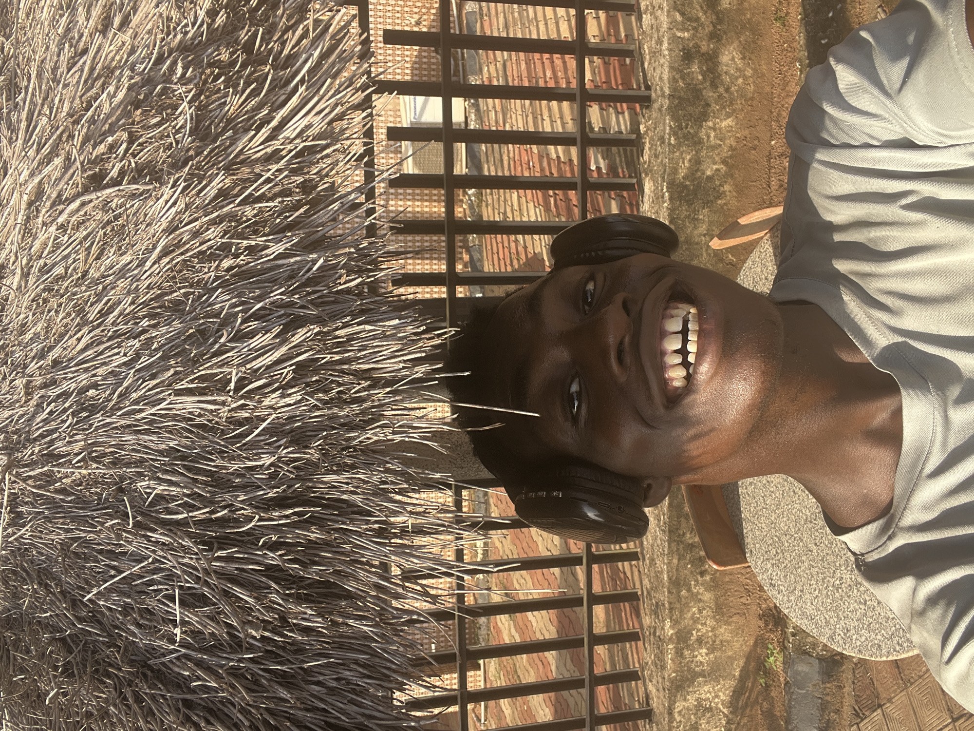 A young, dark-skinned man is captured in a vertical, brightly lit outdoor setting, likely in Nkpor, Nigeria. He is smiling broadly, revealing his teeth, and looking directly at the camera, conveying a cheerful and happy emotion. He wears a light-colored t-shirt and black over-ear headphones.
To the left of the frame, a large mass of dried, thin branches, possibly part of a thatched structure or a decorative element, forms a dense, textured backdrop. Behind the man, a dark metal fence with horizontal bars provides a visual separation from the background. Through the fence, various elements of buildings are visible, including patterned brickwork or tiled roofs, and a sign faintly displaying the word "Panasonic." The ground around the man appears to be paved with light-colored concrete or tiles, and a wooden chair is partially visible to his right.
The scene is illuminated by strong, direct sunlight, creating distinct shadows and indicating a clear, sunny day, likely during mid-morning or afternoon. Text visible on the headphones includes "VOL +/-", a Bluetooth symbol, a power symbol, and "TW 104 1".