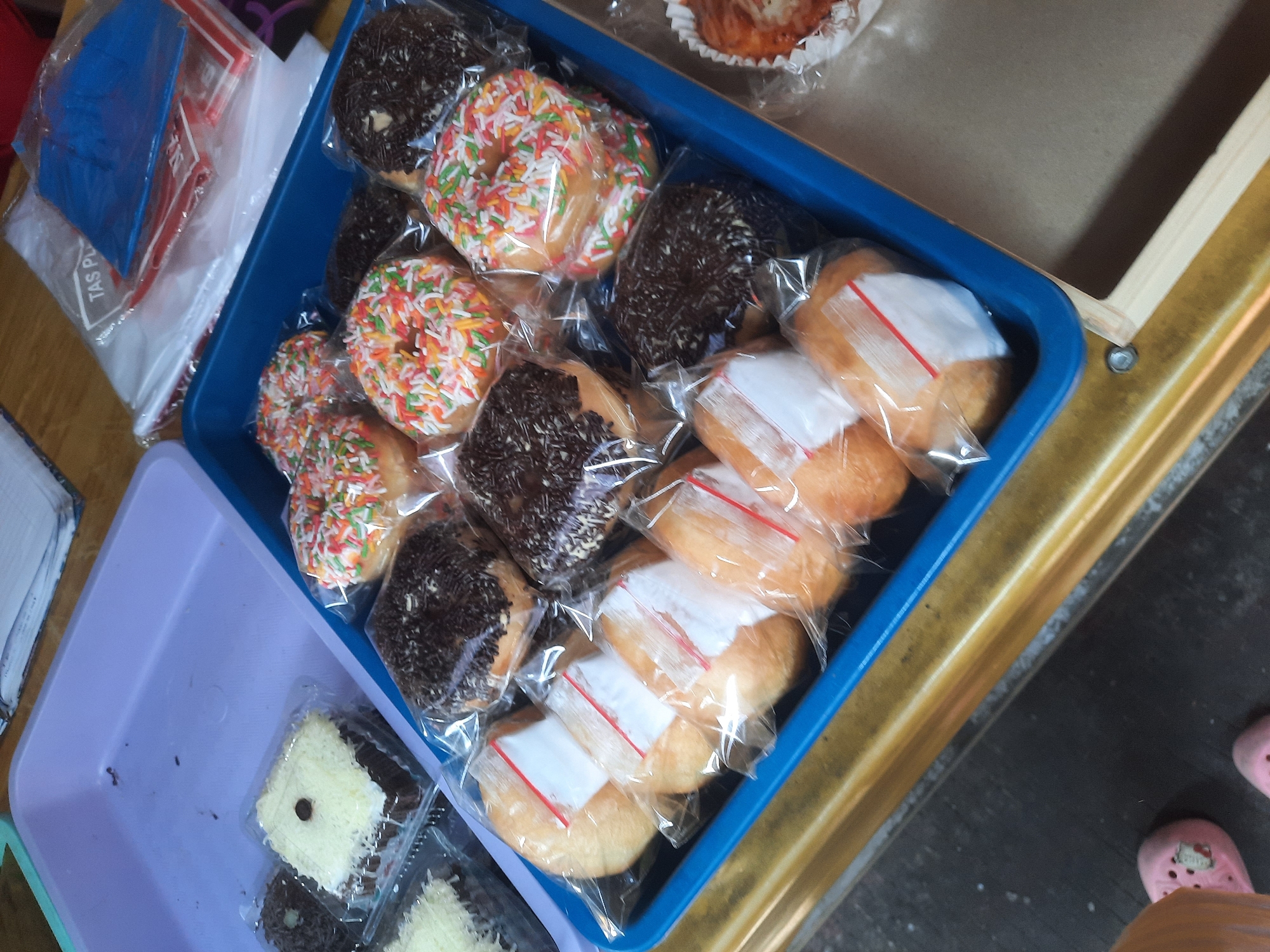 A close-up, high-angle shot shows a blue plastic tray filled with various pastries and snacks. The tray is positioned on a wooden surface, possibly a market stall, with a lighter-colored plastic tray visible in the upper left corner. In the background, several plastic bags with red and blue contents are stacked. The main focus is the blue tray, which contains:
* Several donuts covered in colorful sprinkles.
* Several donuts covered in chocolate sprinkles.
* Several bread rolls, each individually wrapped in clear plastic with a white label.
* A few chocolate cakes topped with white grated cheese and a single chocolate button, also individually packaged in clear plastic.
* In the far right of the tray, a single pastry in a white paper cup, topped with a melted, golden-brown substance and what appears to be a sprinkle of green herbs.
The lighting suggests an indoor or shaded outdoor environment. The bottom left of the image shows a small portion of two pink Crocs-style shoes, indicating a person is present, likely the photographer or a customer. The scene is likely from North Jakarta, Indonesia, given the common types of pastries and the packaging style.