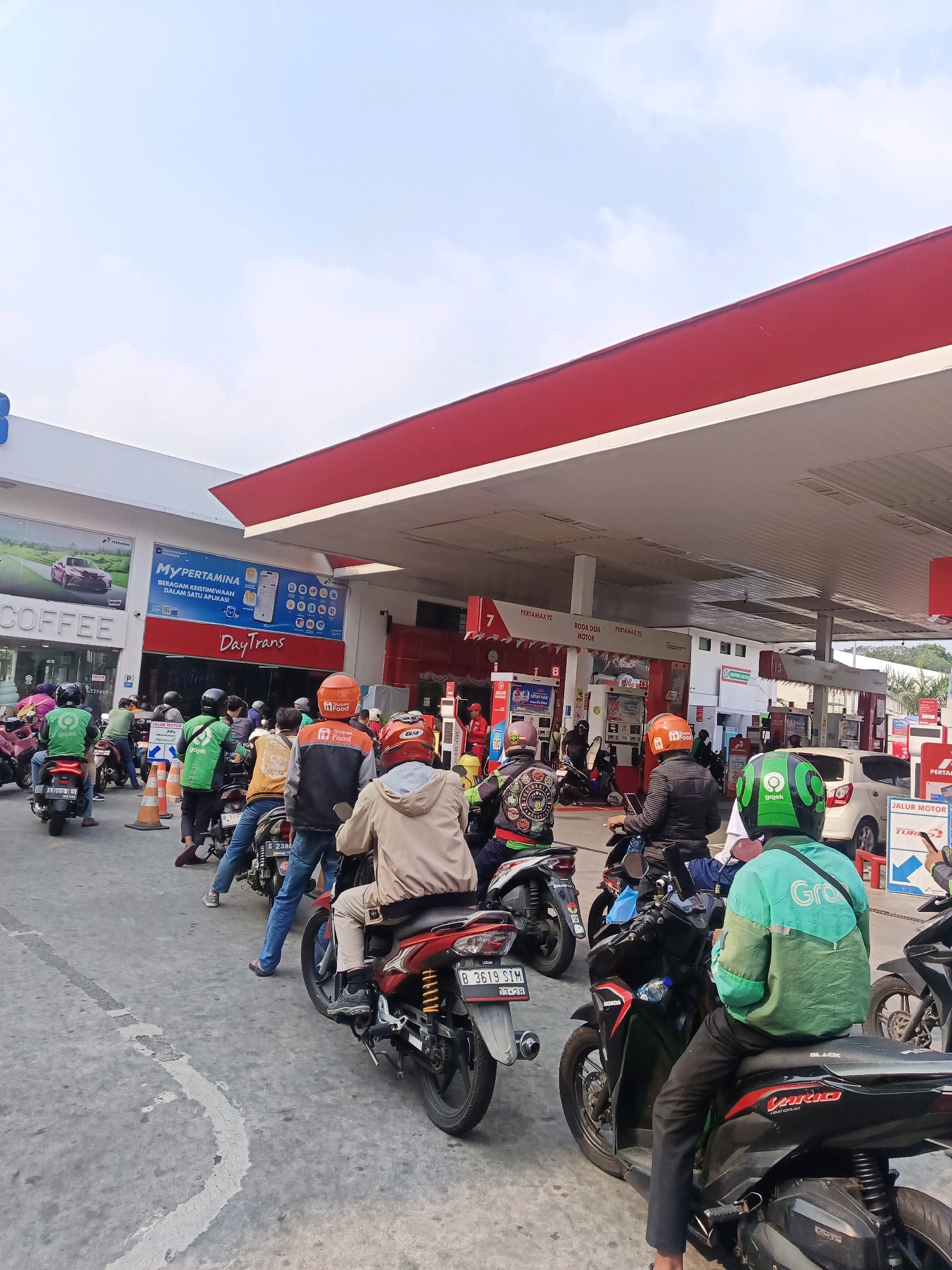 The image captures a queue of motorcyclists at a Pertamina gas station in South Jakarta, Indonesia, under a partly cloudy sky. The foreground features several motorcycles and their riders, many wearing helmets and jackets associated with ride-sharing services like Gojek and Grab. One rider in a green Gojek jacket is prominent, sitting on a black Honda Vario. To the left, a motorcycle with a license plate that reads "B 3619 SIM" is visible. Further back, a line of motorcycles and riders extends towards the gas pumps. The gas station canopy, a prominent red and white structure, extends over the pumps. Signs indicate different fuel types, such as "Pertamax 92." Buildings, including a "Coffee" shop and an area with "DayTrans" branding, are visible behind the queue. The overall scene depicts a common urban scenario of a fuel shortage or high demand at a gas station.
