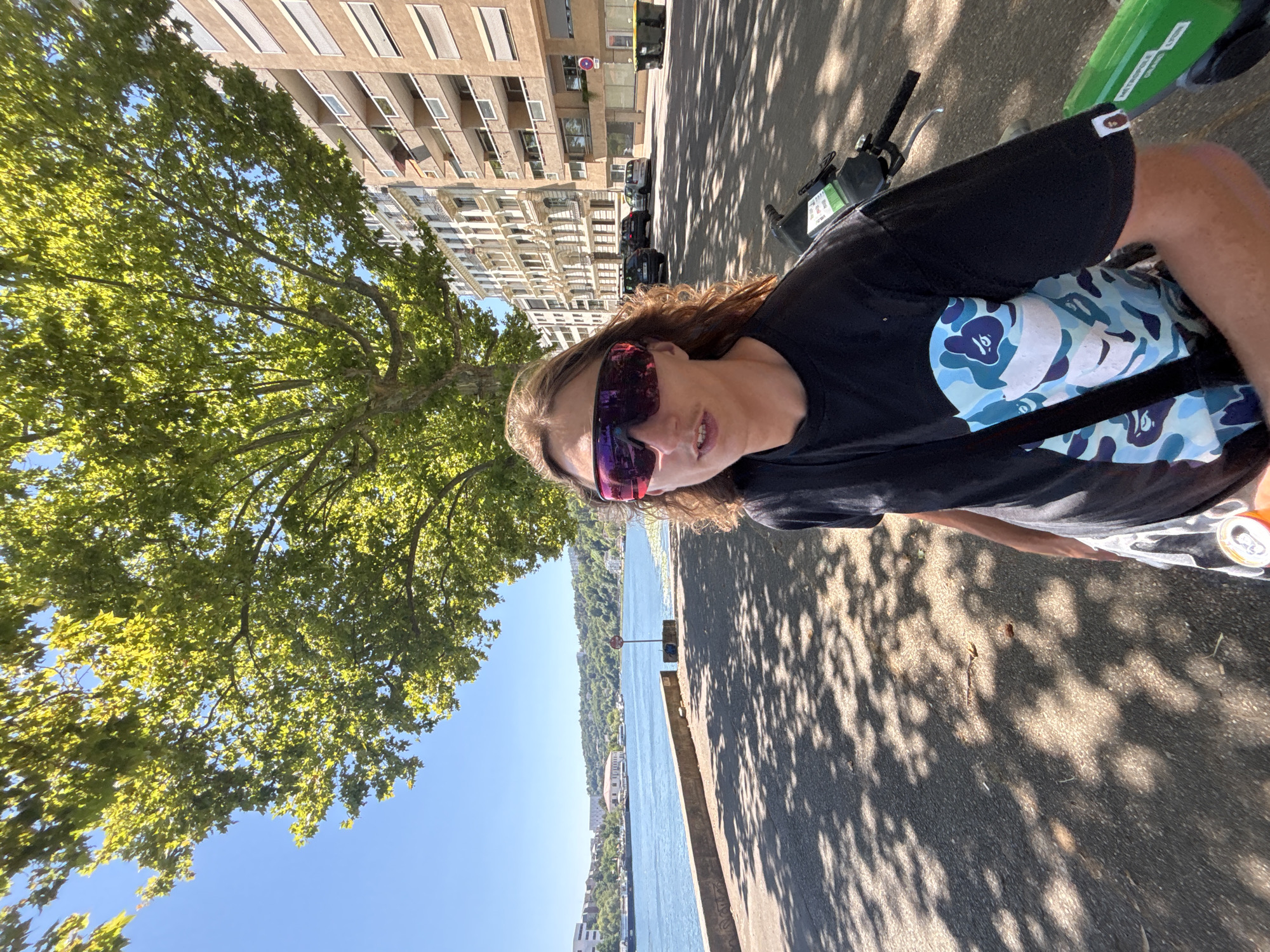 A person with long, brown, wavy hair and sunglasses with pink lenses is standing on a paved area next to a body of water. The person is wearing a black t-shirt with a blue and white camouflage pattern featuring ape heads. Behind the person, there is a tree with green leaves and a street with buildings and parked cars. The body of water appears to be a river or canal, with buildings and greenery on the opposite bank. The scene is illuminated by bright sunlight, casting shadows on the ground. The location is identified as Lyon, France, likely along the Rhône River. The presence of a green public transport scooter suggests an urban setting.