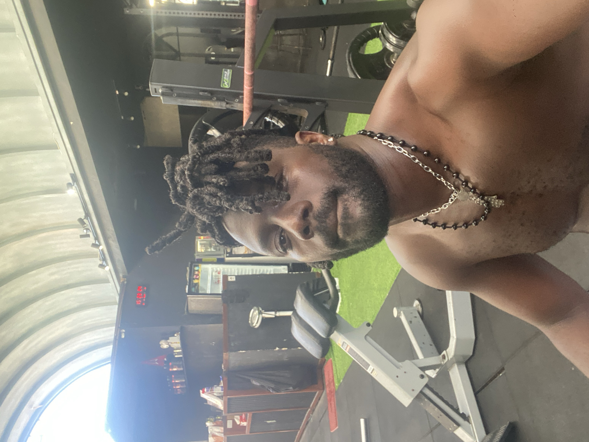 A man with dark skin, dreadlocks, and a beard is taking a selfie in a gym. He is shirtless and wearing two necklaces. In the background, there is gym equipment, including a weight rack and benches. There are also some shelves with various items and a mini-fridge. The gym has a curved ceiling and artificial turf flooring. The image is taken in North Kuta, Indonesia.