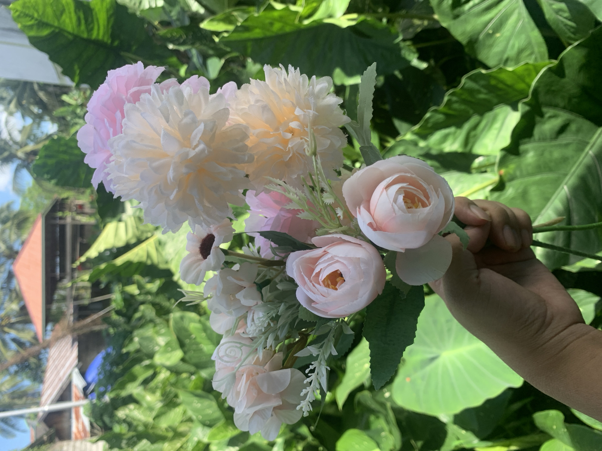 A hand holds a bouquet of artificial flowers, predominantly in shades of pink and white, with a backdrop of lush green foliage and distant buildings. The bouquet consists of several rose-like blossoms and larger, pom-pom style flowers, interspersed with delicate green sprigs. The background reveals tropical vegetation, including large, waxy leaves, palm fronds, and a bright blue sky. In the distance, structures with red and corrugated metal roofs are visible, suggesting a residential or semi-urban setting, potentially in a tropical climate like Kuala Pembuang, Indonesia. The lighting indicates it is daytime, with sunlight illuminating the scene.