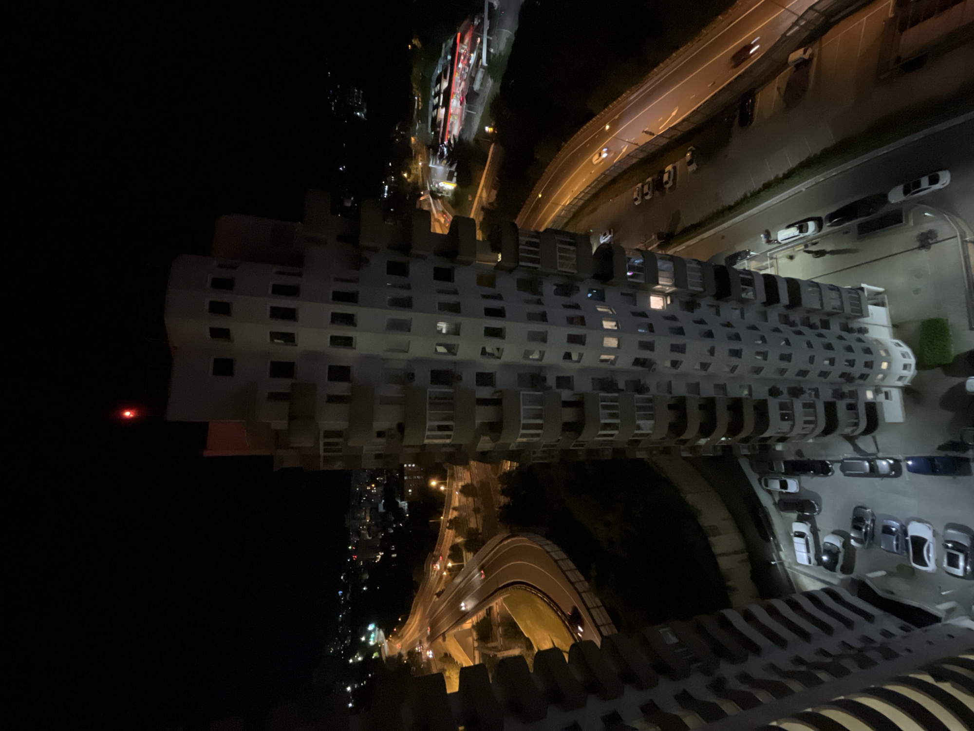 The image depicts a nighttime aerial view of a cityscape in Sochi, Russia. The foreground is dominated by a tall, cylindrical residential building with numerous balconies. To the left of this building, another tall residential structure is partially visible. Below and to the left of the main building, a multi-lane highway curves through the scene, with several cars visible on it. Streetlights illuminate the highway and surrounding areas. Further in the background, more city lights and buildings can be seen, indicating an urban environment. At the very top of the main building in the center of the image, a red light is visible, likely an aviation warning light. The overall impression is one of an urban area at night with a focus on modern architecture and infrastructure.