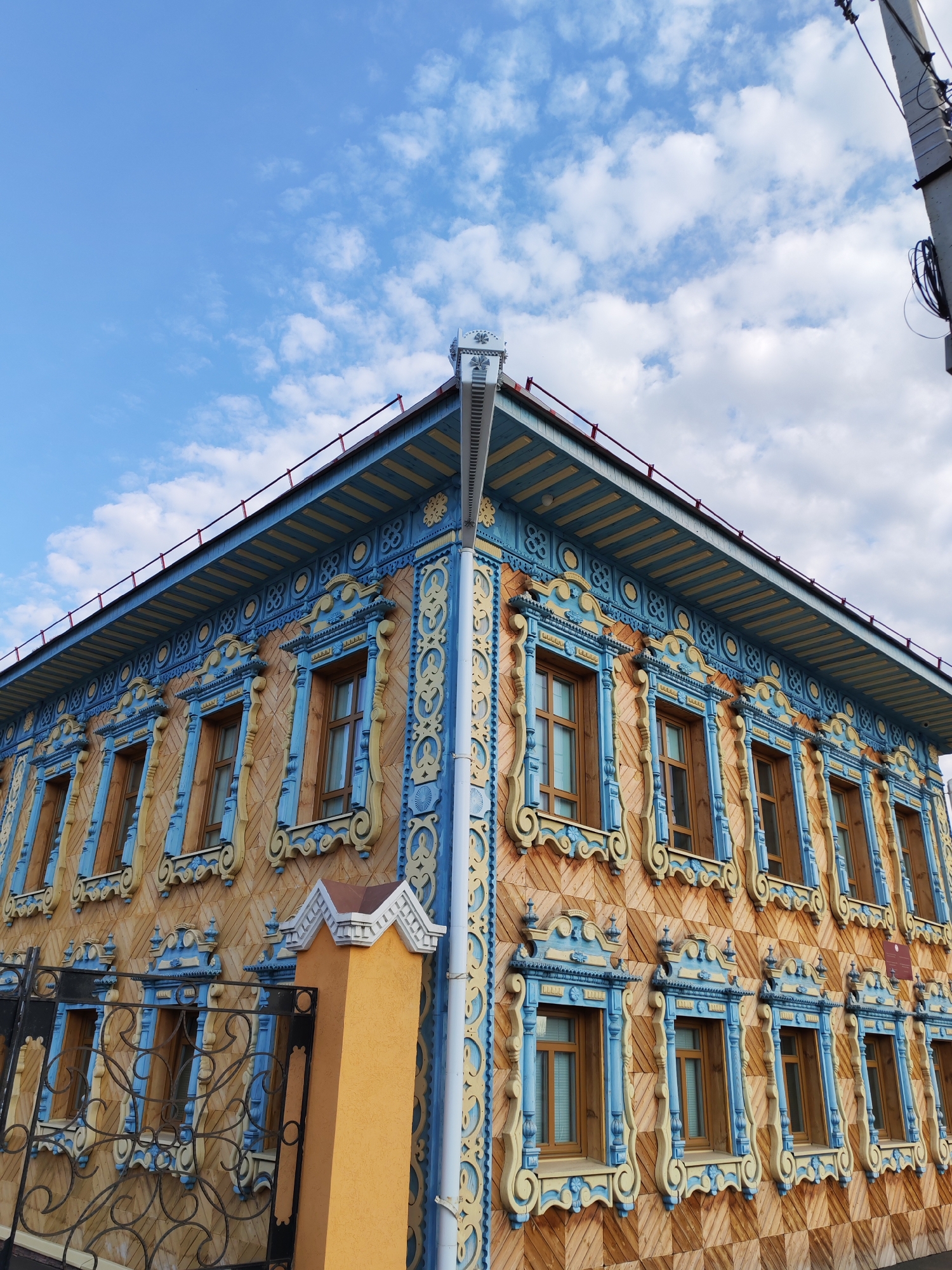 A low-angle, eye-level shot depicts the corner of a two-story building in Sarapul, Russia, framed against a sky with scattered clouds. The building exhibits distinctive architectural features: a brown patterned facade, windows with intricately carved blue and yellow frames, and a blue eaves canopy. A white downspout is attached to the corner of the building. A fence and a yellow pillar stand in front of the building. An electricity pole with wires is in the upper right corner of the scene.