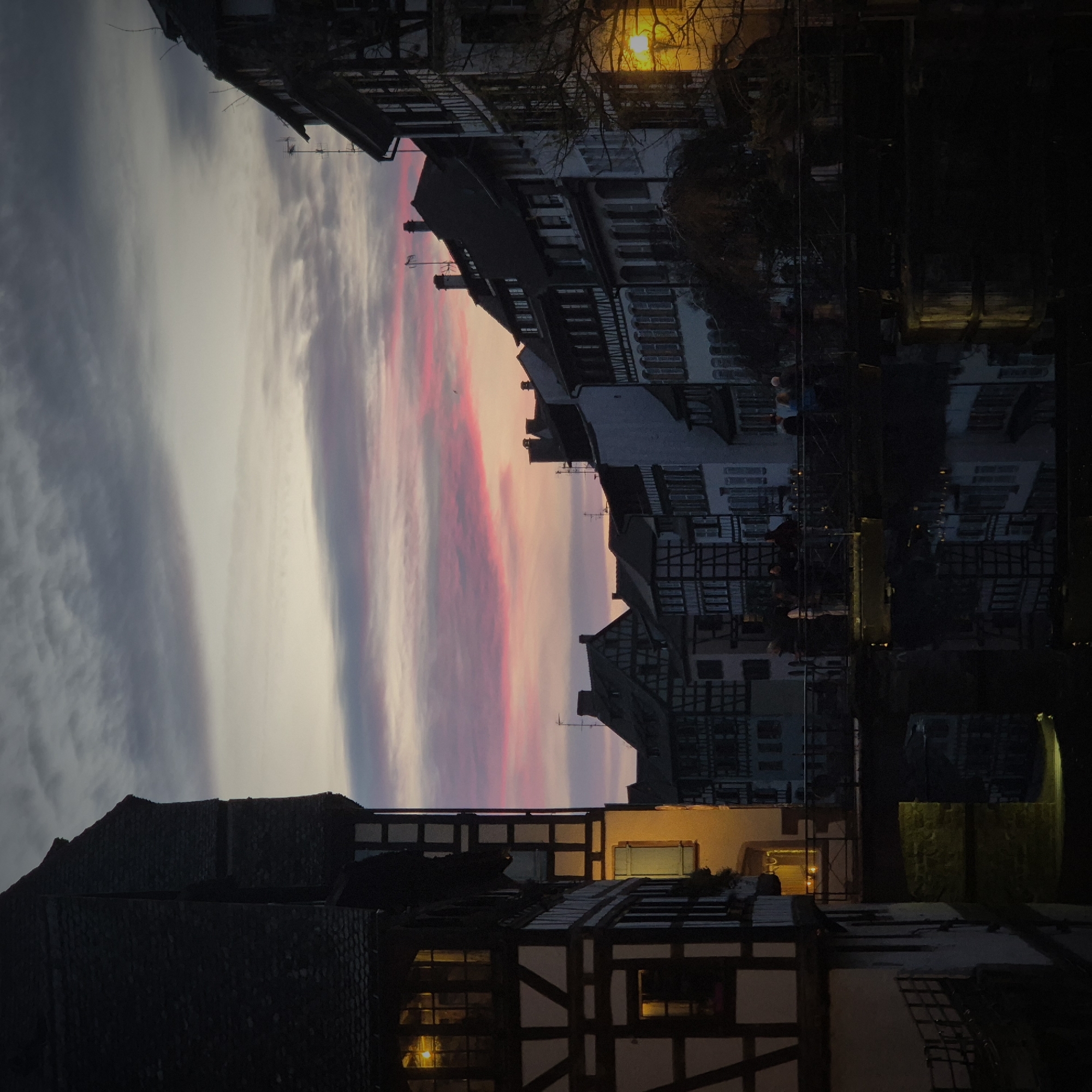 The image depicts a twilight scene in La Petite France, Strasbourg, France, featuring historic half-timbered buildings along a canal. The sky exhibits a gradient of gray and white clouds transitioning to a vibrant band of pink and purple hues near the horizon.
On the left, a multi-story half-timbered building with a dark, steep gable roof dominates the foreground. Its white plastered walls contrast with dark exposed timber beams. Several windows on this building emit a warm yellow light.
A dark canal occupies the lower portion of the scene, reflecting the illuminated building facades and artificial lights. A low bridge with dark railings spans the canal in the middle ground. Approximately ten to fifteen individuals are present on this bridge, mostly appearing as dark silhouettes. Some appear to be standing, while others are in motion.
Along the right bank of the canal and extending into the background, a row of additional half-timbered and plastered buildings with varying rooflines is visible. Many of these buildings also feature dark timber frames and windows, some of which are lit. A prominent yellow streetlight on the far right illuminates the bare branches of a nearby tree. Reflections of building lights are distinctly visible on the water's surface beneath the bridge and in the foreground. The overall scene is dimly lit, relying on the ambient twilight and artificial light sources.
