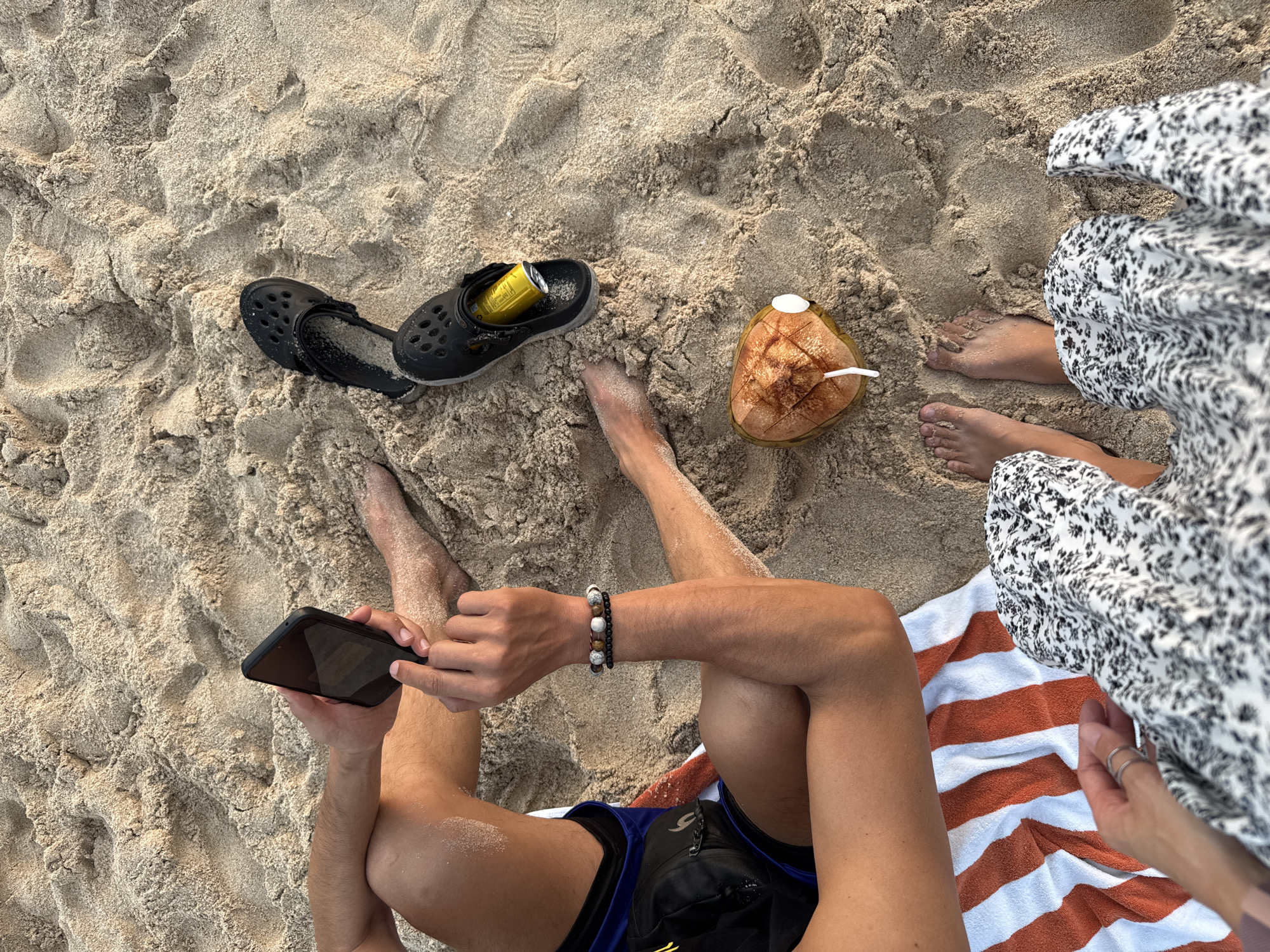 This image depicts a beach scene in Kuta Selatan, Indonesia, featuring two individuals relaxing on the sand. A person on the left, wearing blue and black shorts, is holding a black smartphone. Beside them, a pair of black Crocs are placed on the sand, with one shoe containing a yellow can. A halved coconut with a straw sits in the sand nearby. On the right, another person is dressed in a black and white floral patterned dress. Both individuals' feet are visible, suggesting they are seated or lying down on a striped orange and white towel. The environment is sandy, indicative of a beach, with subtle indentations and ripples in the sand.