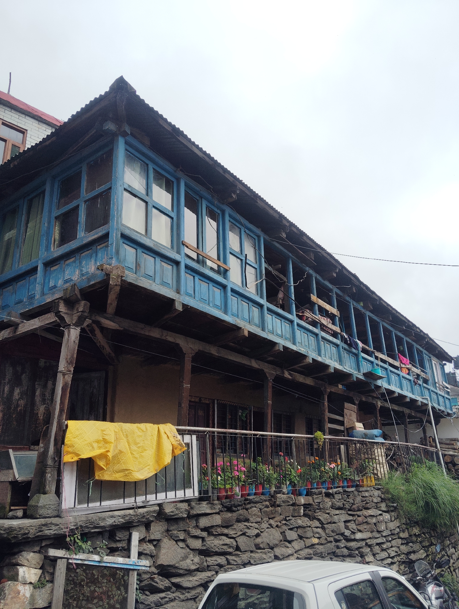 This is a photograph taken in Vashisht, India, showing a traditional wooden building with a prominent blue-painted upper story. The building features a long balcony on the upper level, supported by wooden pillars. The balcony is partially enclosed by wooden railings and has multiple windows on the floor above. Clothes are seen hanging on the balcony railing, and various plants in colorful pots are arranged along the lower railing, in front of a stone wall. A bright yellow tarp is draped over a railing on the lower level. In the foreground, the roof of a white car is visible, and behind it, a motorcycle is parked. The sky is overcast, suggesting a cloudy day. The architecture and the surrounding stone wall indicate a mountainous or hilly region.