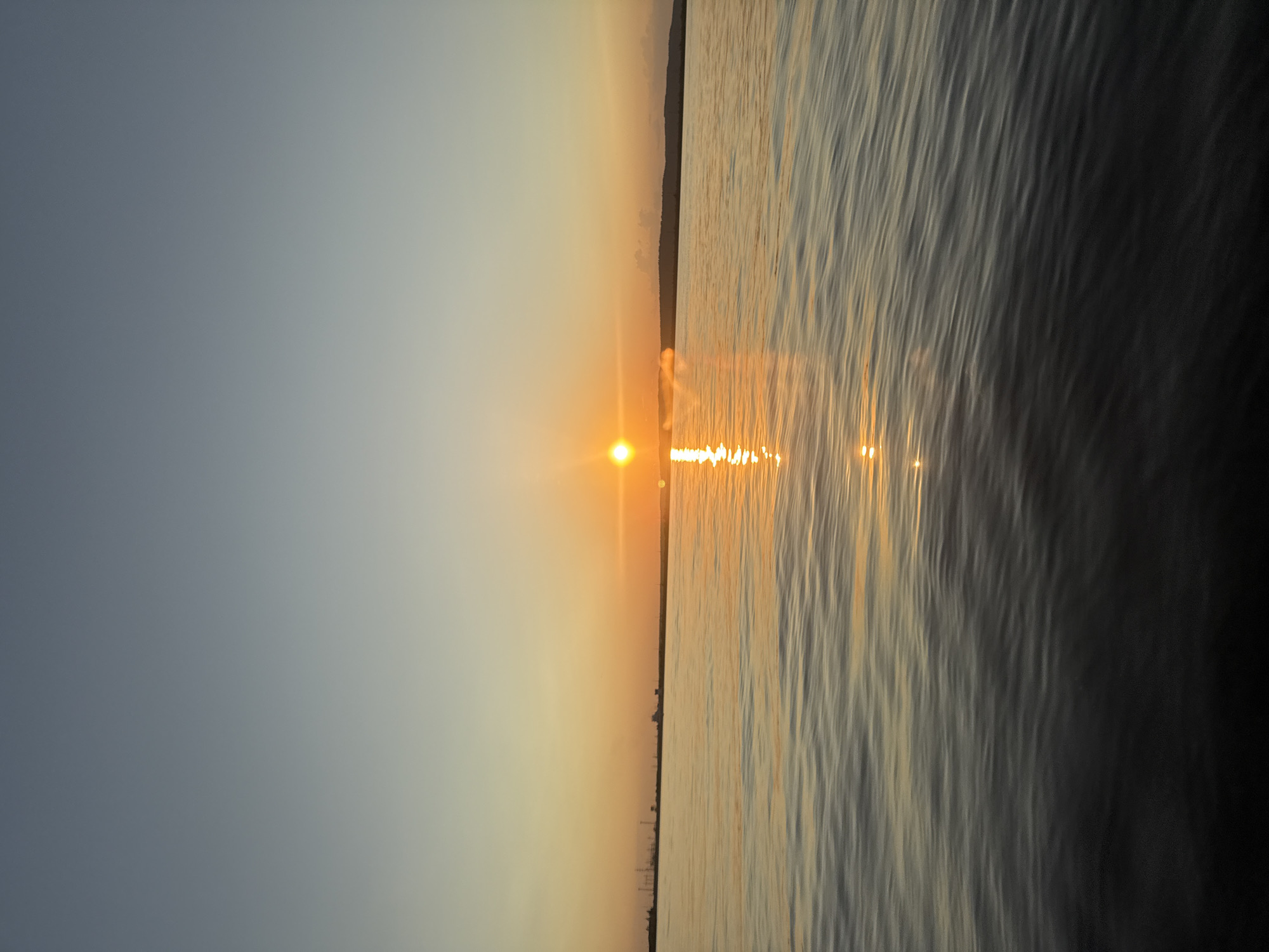 The image depicts a tranquil scene at sunset over a body of water, likely the Kama River, near Chaikovsky, Russia. The sky transitions from a pale blue at the top to a warm orange hue near the horizon. A bright, round sun is positioned slightly left of center, casting a shimmering reflection on the rippling water. In the distance, the silhouette of the Chaikovsky landscape is visible along the horizon, characterized by industrial structures and a gentle, rolling terrain. The foreground is dominated by the dark, textured surface of the water, reflecting the ambient light and colors of the sky. There are no people or specific actions captured in the frame.