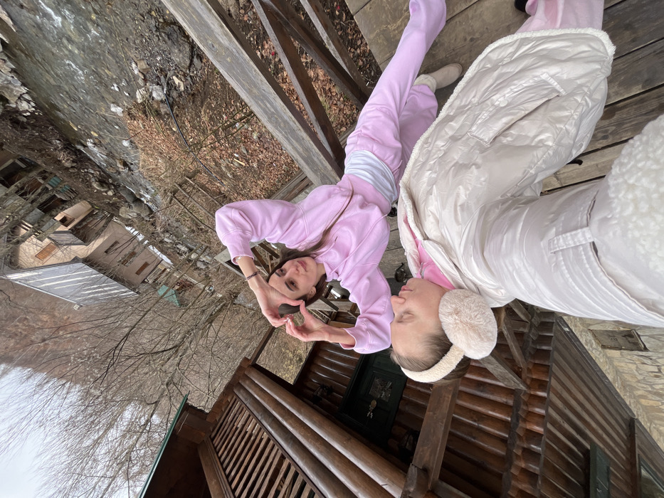 The image captures a high-angle, selfie-style perspective taken on a wooden structure, likely a porch or bridge, located in Crkvine, Montenegro. In the foreground, partially visible, is an individual wearing a light-colored puffer jacket and fluffy cream-colored earmuffs, appearing to hold the camera. Their eyes are closed or looking downwards. Behind this individual, a second female with long dark hair stands, wearing a pink hooded tracksuit over a white top. She is engaged in the action of forming a heart shape with her hands, extending her arms outwards, and looking towards the right of the frame. The immediate environment includes a rough-hewn wooden wall to the left, and visible wooden planks and railings of the structure. The background reveals a narrow, rocky river or stream flowing below. Across the river, multiple rustic buildings with varying rooflines and wooden balconies are visible amidst sparse, bare deciduous trees and dry brown foliage, characteristic of a colder season. Beyond the river and buildings, the landscape consists of steep, deforested mountainous slopes under a uniform, overcast sky.