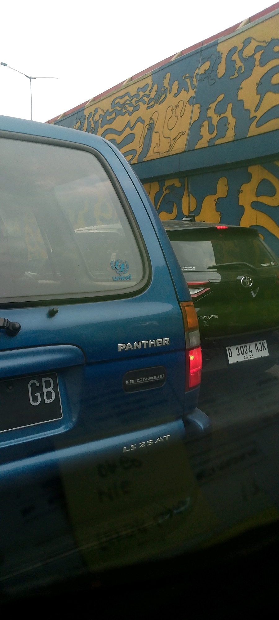 A medium shot captures the rear of a blue minivan with a "PANTHER" emblem, "HI GRADE" designation, and "LS 25AT" model name. The minivan has a license plate with the characters "GB". To the right, the rear of a black SUV is partially visible with a license plate that reads "D 1024 AJK" and a Toyota logo. Both vehicles appear to be in traffic, suggested by the presence of another vehicle behind them and a mural on a structure above. The mural is abstract, featuring yellow and blue shapes and figures. A street lamp is visible in the upper left corner of the frame. The overall scene appears to be in an urban setting, possibly during daylight, given the lighting conditions. The presence of the license plate "D" suggests the location is in Java, Indonesia.