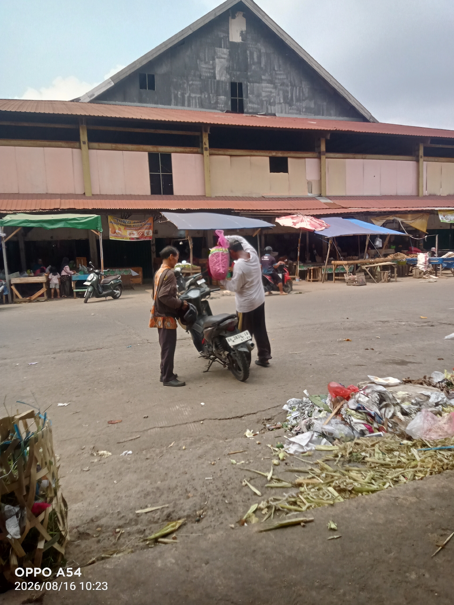 In Jambi, Indonesia, a market scene unfolds under an overcast sky. Two men stand beside a parked motorcycle in the foreground. The man on the left, wearing a brown jacket and an orange-patterned shirt, holds a black helmet. The man on the right, in a light-colored long-sleeved shirt and dark pants, carries a pink plastic bag. Behind them, a market building with a corrugated metal roof looms, its facade a mix of beige and pink panels. Stalls with various awnings, some green, red, and blue, line the street, displaying goods. In the background, other market-goers are visible, some interacting with vendors. The street is paved with asphalt and shows signs of wear and tear. To the right of the main subjects, a large pile of trash, including plastic bags, wrappers, and organic waste, accumulates on the ground, with a woven basket nearby also filled with debris. The timestamp on the image indicates it was captured on August 16, 2026, at 10:23 AM.