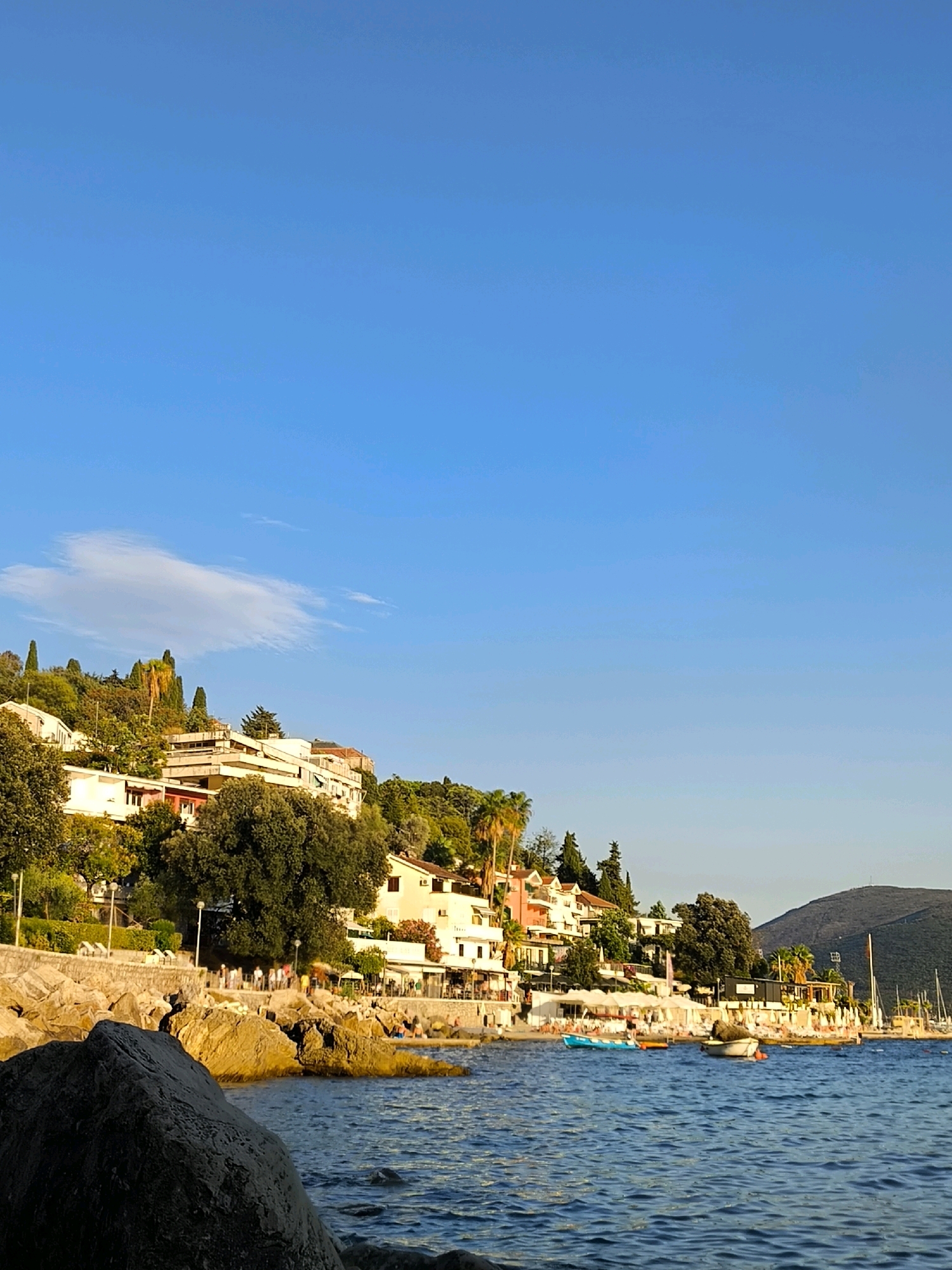 The image captures a coastal scene in Herceg Novi, Montenegro, likely during late afternoon or early evening due to the warm, angled sunlight. The foreground features a large, rough-textured rock, casting a shadow into the clear blue water. The water itself is calm, with gentle ripples reflecting the bright sky.
Along the coastline, a stone embankment separates the water from a promenade lined with buildings. These buildings are a mix of multi-story structures, some appearing to be hotels or apartments, with white and cream facades, interspersed with red-tiled roofs. They are nestled on a lush green hillside dotted with various trees, including tall cypress trees and palm trees. Small umbrellas and beach chairs are visible on the shore, suggesting a resort or beach area.
Several small boats are moored or floating on the water, including a small blue dinghy and a larger boat with a covered area. Further in the background, a dark, verdant mountain slopes down towards the sea. A few people are visible on the promenade and on the beach area, appearing to be casually strolling or relaxing. The overall atmosphere is serene and picturesque, characteristic of a Mediterranean coastal town.