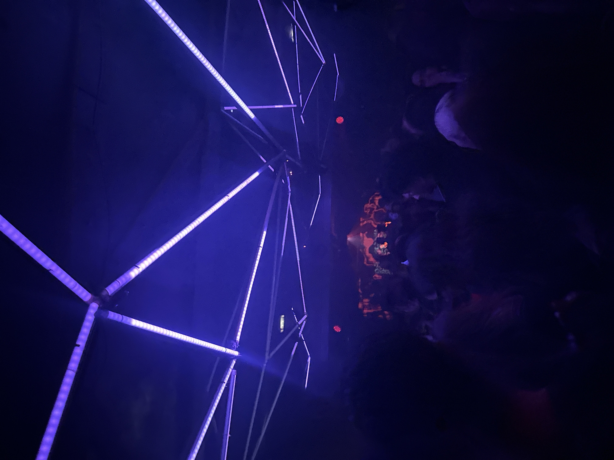 The image displays an indoor scene within a dark, crowded establishment, identified as a nightclub or music venue in Paris, France. The ceiling features a complex geometric array of tubular light fixtures housing numerous purple and blue LED strips, casting a dominant violet illumination across the space. Below this overhead lighting structure, a dense crowd of individuals is visible in the mid-ground and background. Most people appear as indistinct dark silhouettes, illuminated primarily by the ambient purple light, with some areas of the crowd showing faint reddish hues. Individuals are standing in close proximity, suggesting a social gathering. Specific actions or interactions between people are not discernible due to the low light conditions and distance from the viewer.