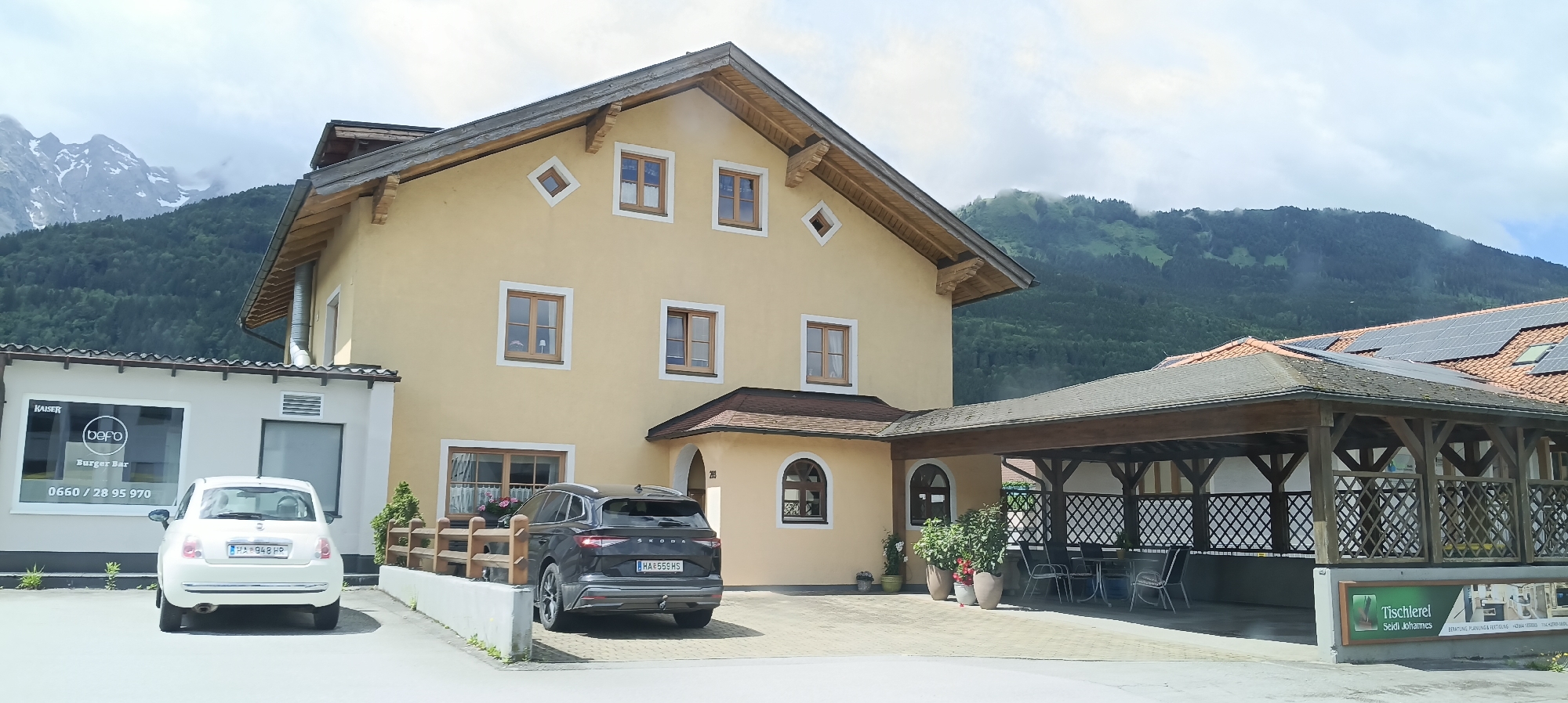 The image shows a building in Moos-Siedlung, Austria, with a light yellow facade and a brown, gabled roof. The building has several square windows with wooden frames and a few diamond-shaped decorative windows. To the left, a smaller white building is visible, featuring a sign with "KAISER bepo Burger Bar" and a phone number. A white Fiat 500 is parked in front of the smaller building. To the right of the main building, a dark grey Skoda Enyaq is parked. Adjacent to the main building is a covered outdoor seating area with a wooden roof supported by posts, furnished with chairs and tables. A sign with "Tischlerei Seidl Johannes" is mounted on a green background at the edge of this covered area. In the background, a lush green mountain slope rises, with a snow-capped peak visible above it. The sky is partly cloudy.