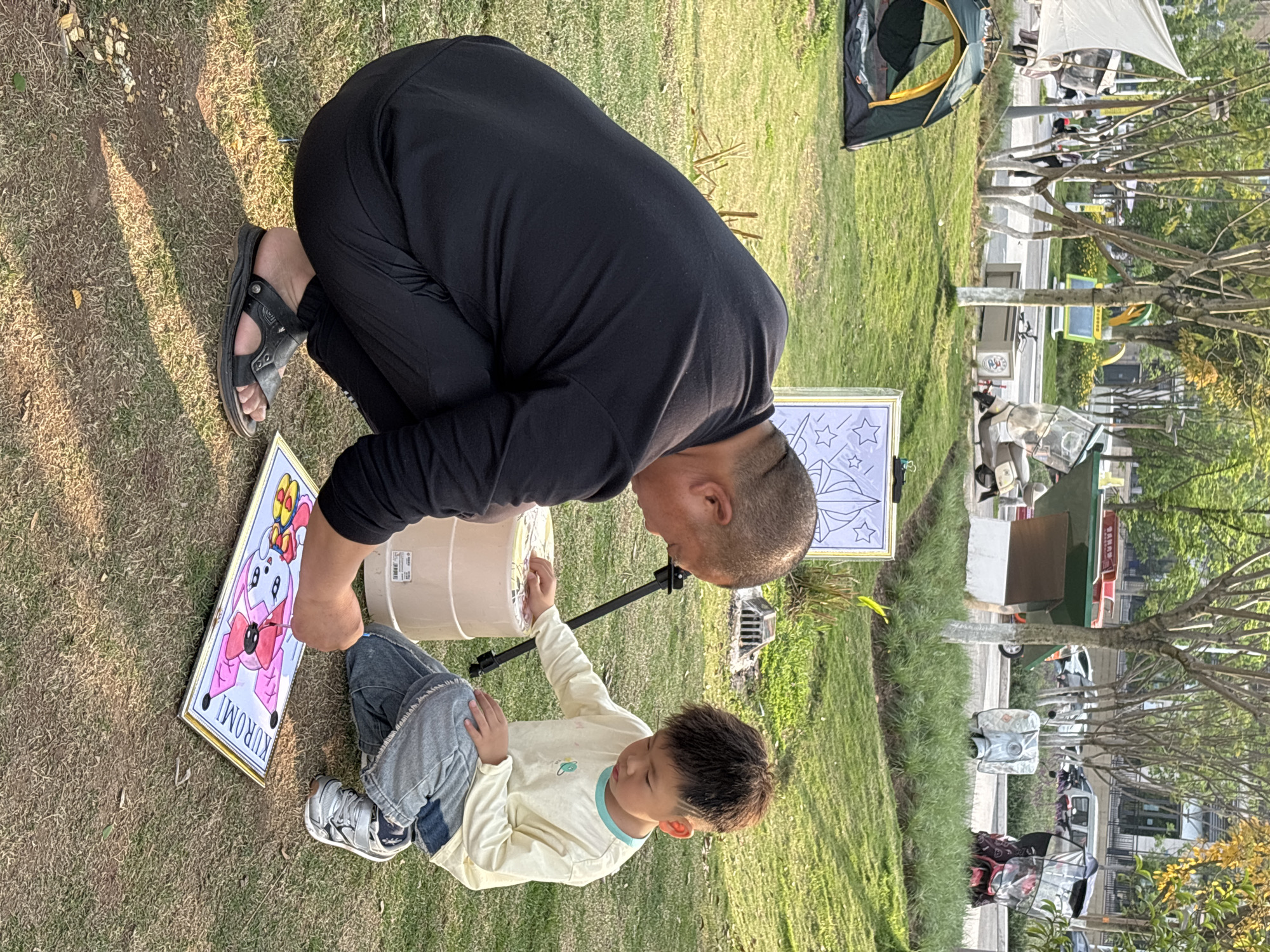 A man and a child are outside on a grassy area. The man is kneeling, dressed in black, wearing sandals. A child kneels beside him, wearing a light-colored long-sleeve shirt and jeans. They appear to be painting or drawing. Next to them are two framed images: one is a character drawing, and the other is a black-and-white drawing of stars. In the background, there is a tent, trees, and buildings, suggesting the scene is in a city, specifically Jiangjin, China.