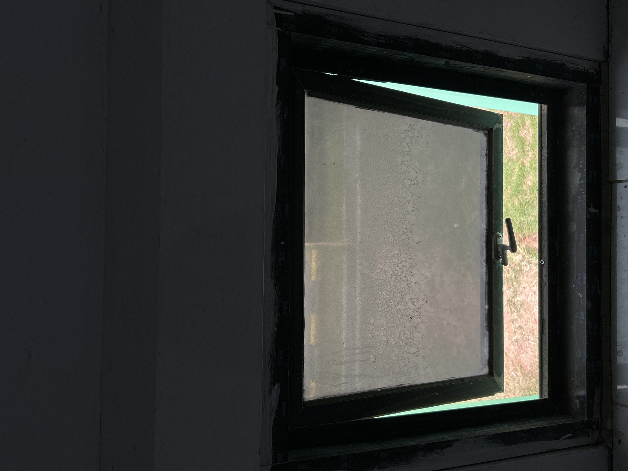 The image shows an interior view looking out through a partially open window in Reuleut Barat, Indonesia. The window frame is dark, likely black or dark brown, and appears to be made of wood or metal, showing signs of wear and possibly paint residue around its edges. The glass pane of the open window is dirty or frosted, exhibiting a distinct speckled pattern across its surface. A metallic window latch or handle is visible on this pane.
Through the open section of the window, a bright outdoor scene is visible, dominated by green grass and patches of reddish-brown earth. The interior space to the left and around the window frame is a dark grey, indicating a dimly lit room or shaded area, contrasting with the bright exterior. The illumination from outside suggests it is daytime. Faint, repeating white text or a logo, possibly "Vilcal" or "Vilca," is discernible on the dark window frame, particularly on the vertical sections adjacent to the wall. No people or specific events are depicted, just the architectural detail and the outdoor view.