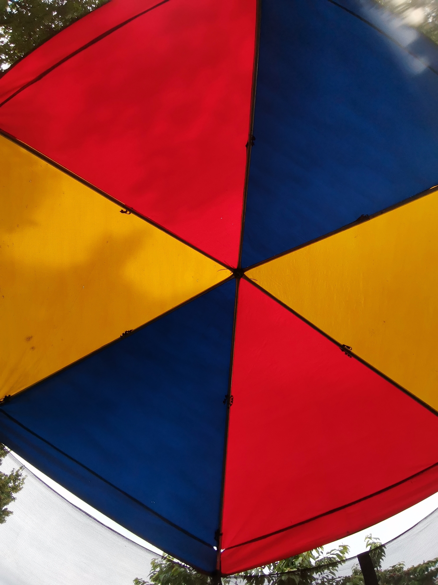 This media file shows a close-up, low-angle view of a colorful outdoor umbrella, likely a canopy or part of a play structure. The umbrella is divided into alternating triangular sections of bright red, yellow, and blue fabric. The geometric pattern is symmetrical, with four main sections radiating from a central point, creating a star-like or pinwheel effect.
The fabric appears slightly textured and shows some minor wear and tear, with small dark clips or fasteners visible along the seams where the fabric panels meet. In the upper portion of the image, glimpses of green foliage from trees can be seen against a bright sky, suggesting it is daytime. The light filtering through the canopy creates subtle variations in the colors.
In the lower part of the frame, a white mesh material is visible, possibly part of a net or enclosure beneath the umbrella. The overall impression is one of an outdoor recreational space, perhaps a park, playground, or garden. The weather appears to be fair, judging by the brightness of the sky and the visible foliage. No people are visible in the frame, and there is no visible text. The setting is identified as Osoitsa, Bulgaria.