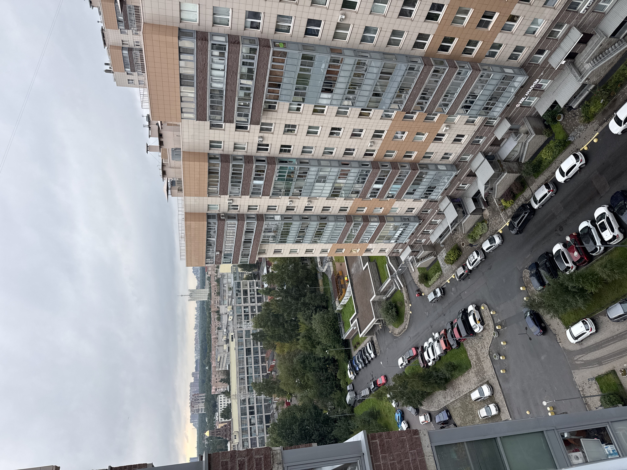 This is a high-angle, exterior, daylight, medium shot of a modern apartment complex in Sinyavino, Russia, on an overcast day. The primary subject is a multi-story, beige and brown brick apartment building with numerous windows and balconies. In the foreground, a brick building, likely another apartment building, occupies the left side of the frame. Below the apartment buildings, a paved courtyard area with a parking lot is visible. Several cars are parked in the lot, including white, black, red, and blue vehicles. A patch of green grass and trees separates the parking lot from the apartment buildings. In the background, other buildings and a densely wooded area can be seen. The sky is filled with gray clouds. The overall scene depicts a residential urban environment.