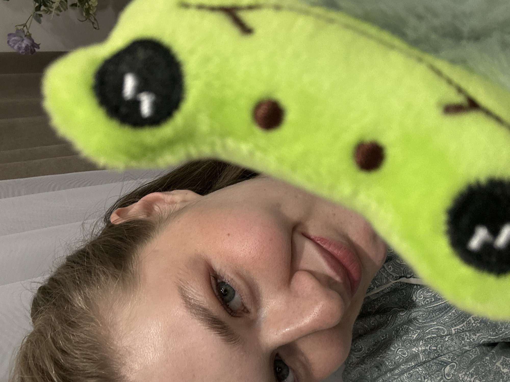 A close-up, low-angle indoor shot presents a woman's face partially obscured by a green plush toy. The woman has fair skin, light brown hair, and blue-green eyes. Her lips are pink, and the details of her nose and cheek are visible. The green plush toy features black eyes with white highlights and small brown dots. The background includes a white, possibly sheer fabric, and hints of light-colored stairs. A blurry image of white flowers is visible in the upper left corner. The woman appears to be lying on a patterned surface, possibly bedding. There is no indication of the location being Kazan, Russia.