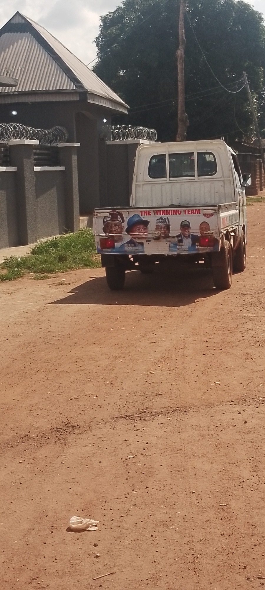 A white pickup truck with a decal on its tailgate reads "THE WINNING TEAM" and displays images of four men in hats. The truck is driving on a dirt road. The scene appears to be outdoors, possibly in a rural or semi-urban area, given the presence of a dirt road, trees in the background, and a building with a corrugated metal roof and barbed wire fencing on the left. The sky is cloudy, suggesting it might be a partly cloudy day. The overall impression is that of a political campaign or advertisement, with the truck likely used for transport or promotional purposes. The image is taken from behind the truck, focusing on its rear and the decal. The ground is composed of reddish-brown dirt with scattered small stones. In the lower left corner of the frame, a crumpled white plastic bag lies on the ground.