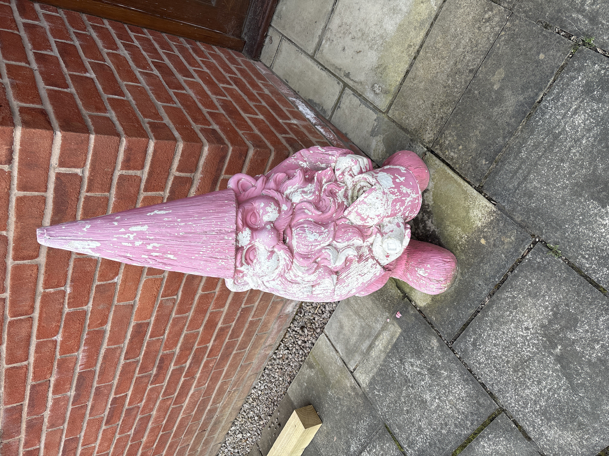 An outdoor ground-level shot shows a pink and white painted statue of an ice cream cone resting against the brick wall of a building in the city of Shrewsbury, United Kingdom. The statue is positioned horizontally in the frame. The cone section points towards the left and the multiple scoops of ice cream extend to the right. The paint is faded and chipped, revealing the base material in places. The wall is constructed of red bricks with light-colored mortar and forms a step or platform alongside the building. Adjacent to the wall is a paved area composed of gray concrete slabs. Between the brick wall and concrete slabs is a narrow strip of gravel, and a piece of wood lies nearby. The image is taken in daylight, and the environment appears to be an exterior location.