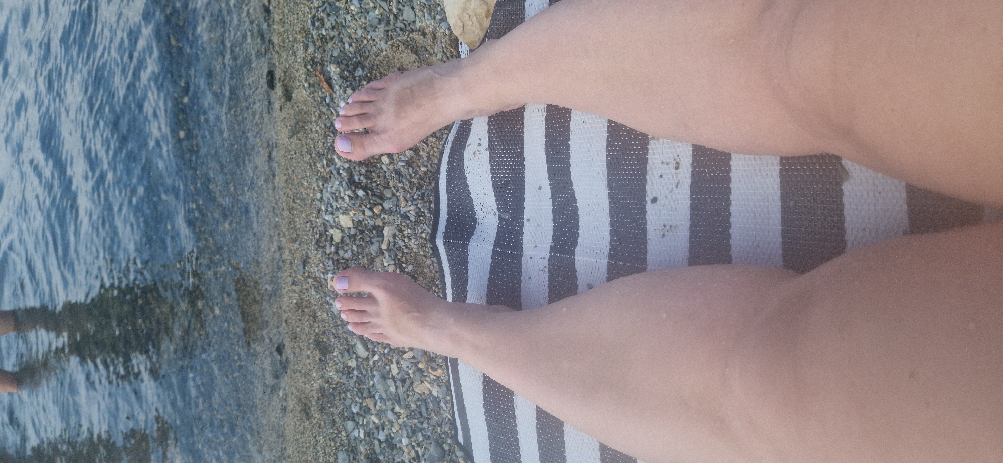 The image shows a pair of bare feet with light pink nail polish resting on a striped mat. The mat is placed on a pebble beach, with the edge of the water visible in the background. The water is calm and appears to be shallow near the shore, with ripples and reflections of light. The feet are positioned centrally in the frame, with the legs extending downwards out of view. The lighting suggests it is daytime, with soft shadows. The overall impression is one of relaxation and a casual beach setting.