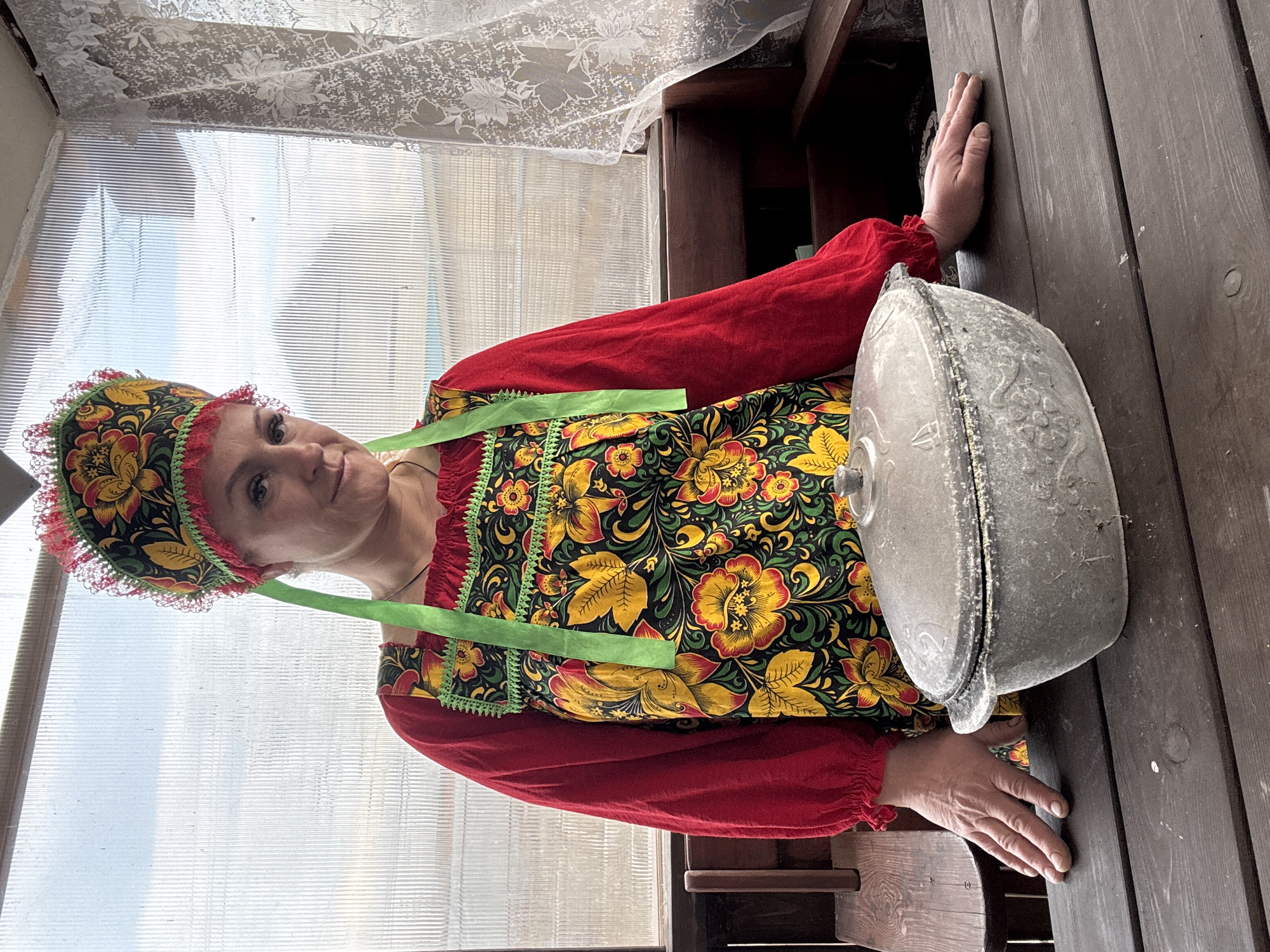 A woman wearing a traditional Russian costume stands beside a table with a heavy, lidded pot on it. She has a floral headdress, a red top with matching cuffs, and a floral apron with green straps. The table is made of dark wood, and the pot appears to be cast iron. The background includes a window with lace curtains. The photograph was taken in Chaikovsky, Russia.