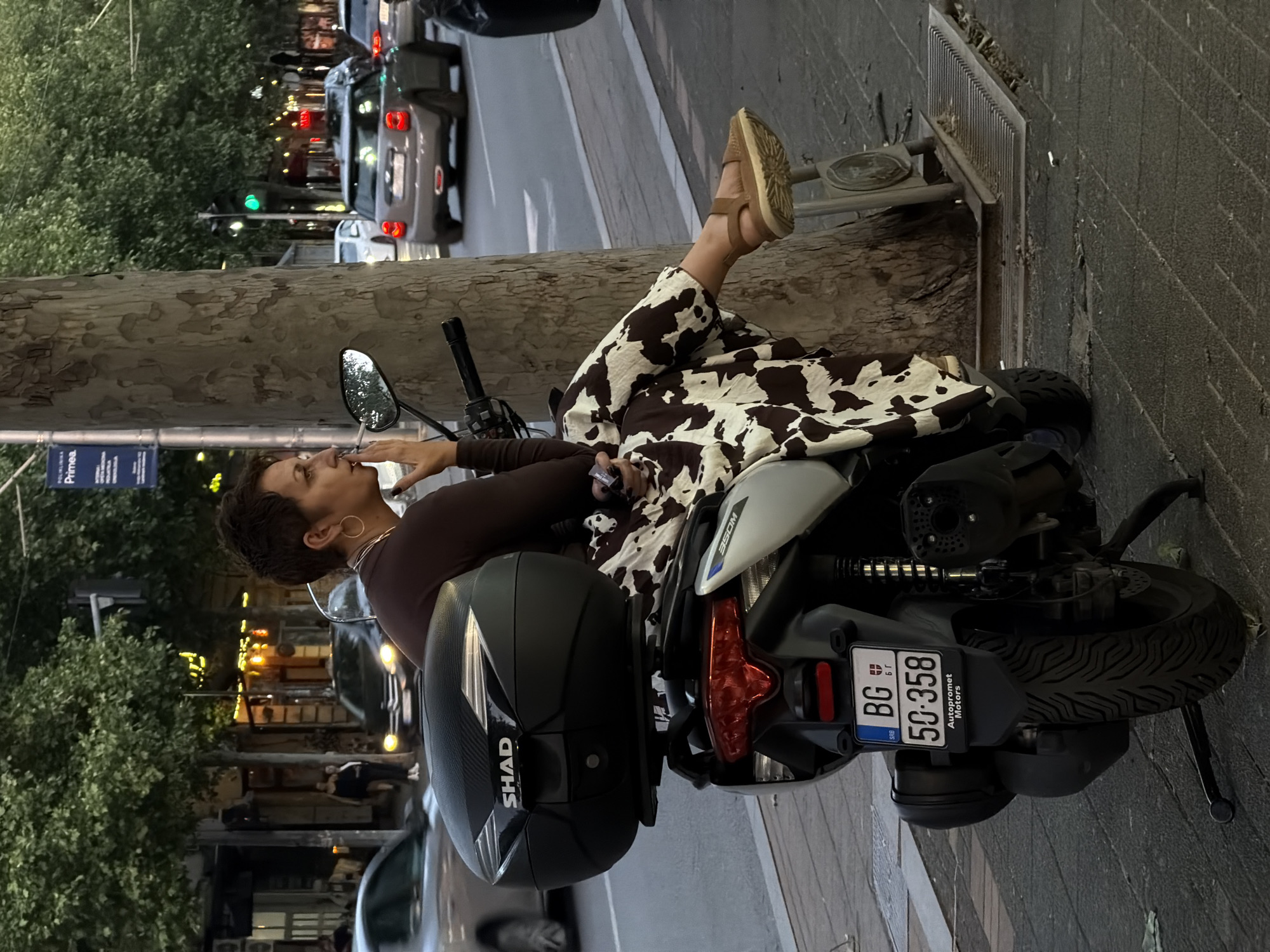 The image shows a woman sitting on a moped with the license plate BG 50-358. She is wearing a brown long-sleeved shirt and cow-print pants. She is smoking a cigarette, and her feet are propped up on a metal grate near a tree. The moped has a black SHAD trunk. The background shows a street in Belgrade, Serbia, with cars and buildings visible.